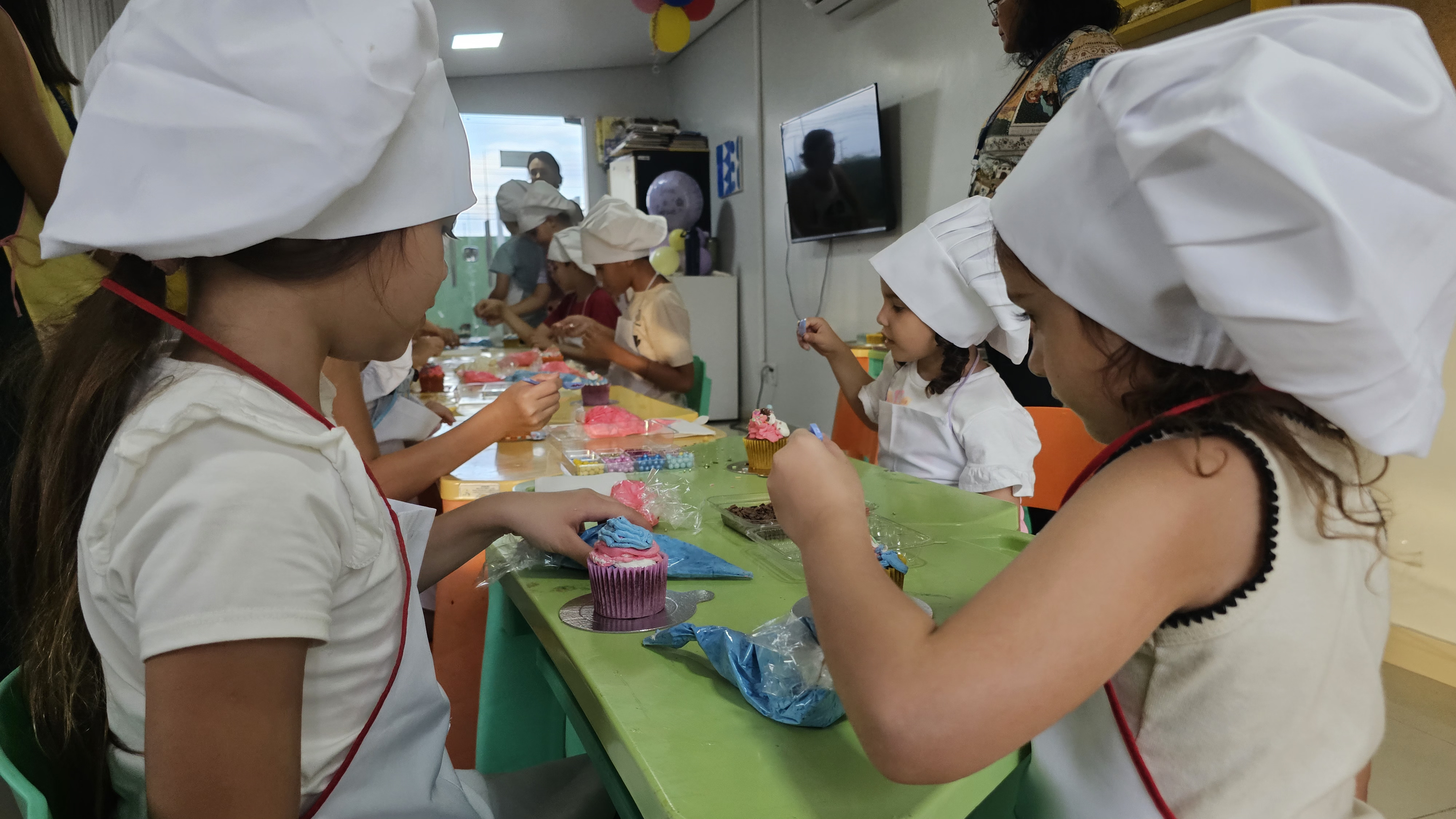 Imagem colorida mostra crianças vestidas de chef, com toucas brancas e aventais, decorando cupcakes coloridos em uma mesa verde. A oficina de culinária acontece em um ambiente interno iluminado com outras pessoas ao fundo. Imagem colorida mostra crianças vestidas de chef, com toucas brancas e aventais, decorando cupcakes coloridos em uma mesa verde. A oficina de culinária acontece em um ambiente interno iluminado com outras pessoas ao fundo.