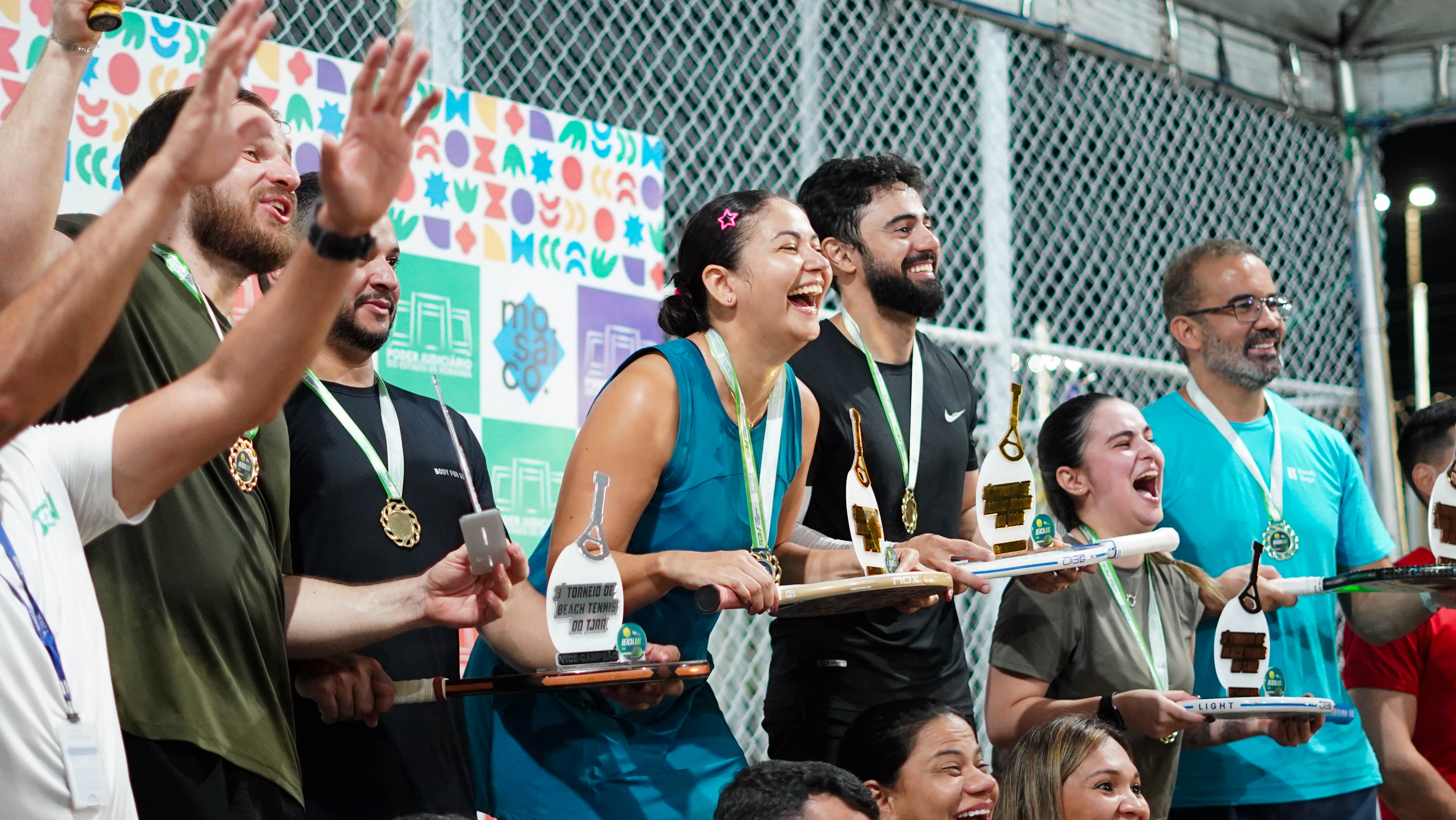 Imagem colorida mostra participantes do torneio durante a entrega de medalhas e troféus. Homens e mulheres aparecem sorrindo, alguns com medalhas no pescoço e troféus nas mãos, diante de um painel colorido montado ao lado da quadra. Imagem colorida mostra participantes do torneio durante a entrega de medalhas e troféus. Homens e mulheres aparecem sorrindo, alguns com medalhas no pescoço e troféus nas mãos, diante de um painel colorido montado ao lado da quadra.