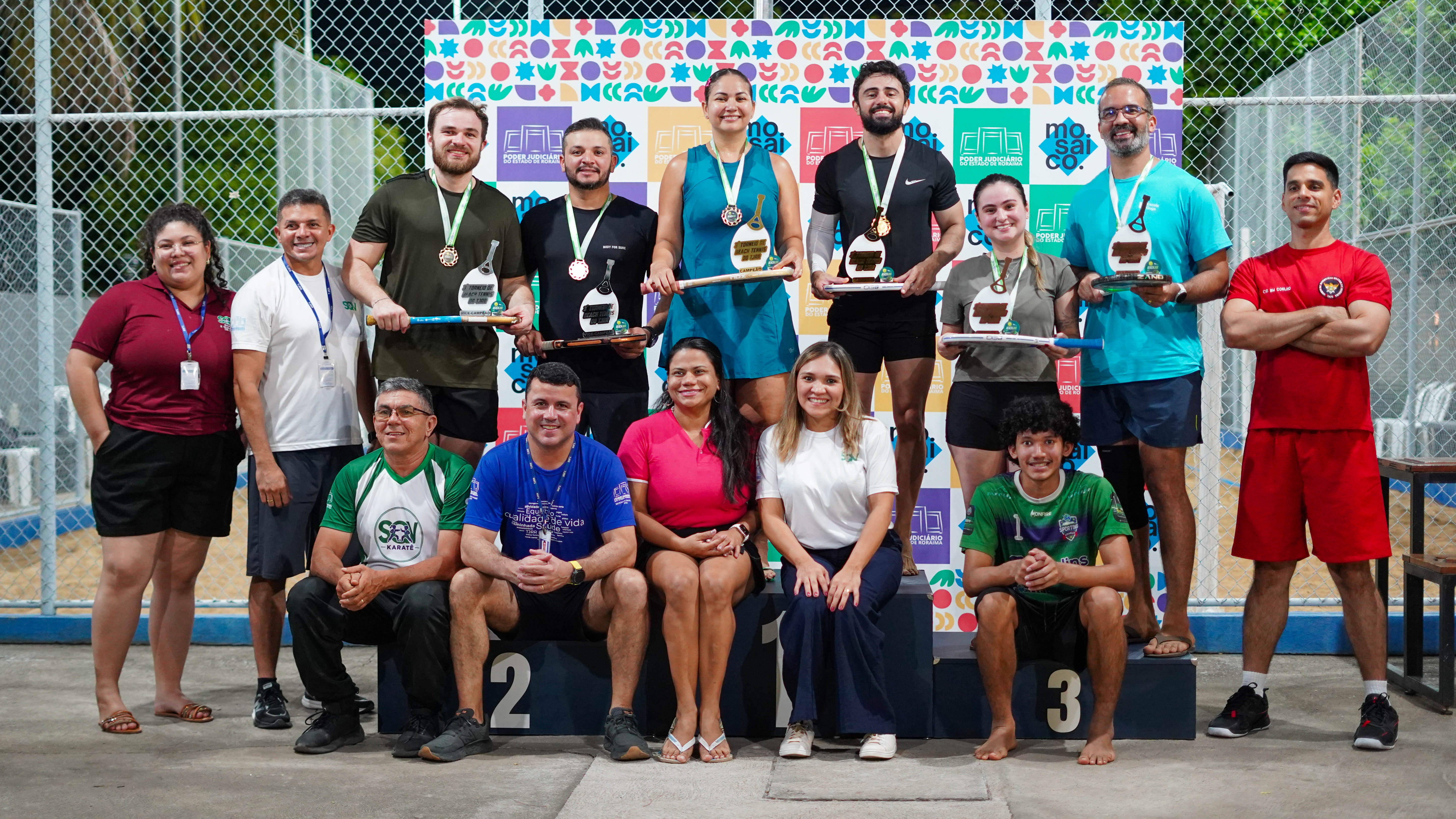 Imagem colorida mostra participantes e integrantes da organização reunidos para uma foto ao final do torneio. Parte do grupo está sobre o pódio, segurando medalhas e troféus, enquanto outras pessoas aparecem à frente e nas laterais. Ao fundo, há um painel colorido com as marcas institucionais. Imagem colorida mostra participantes e integrantes da organização reunidos para uma foto ao final do torneio. Parte do grupo está sobre o pódio, segurando medalhas e troféus, enquanto outras pessoas aparecem à frente e nas laterais. Ao fundo, há um painel colorido com as marcas institucionais.