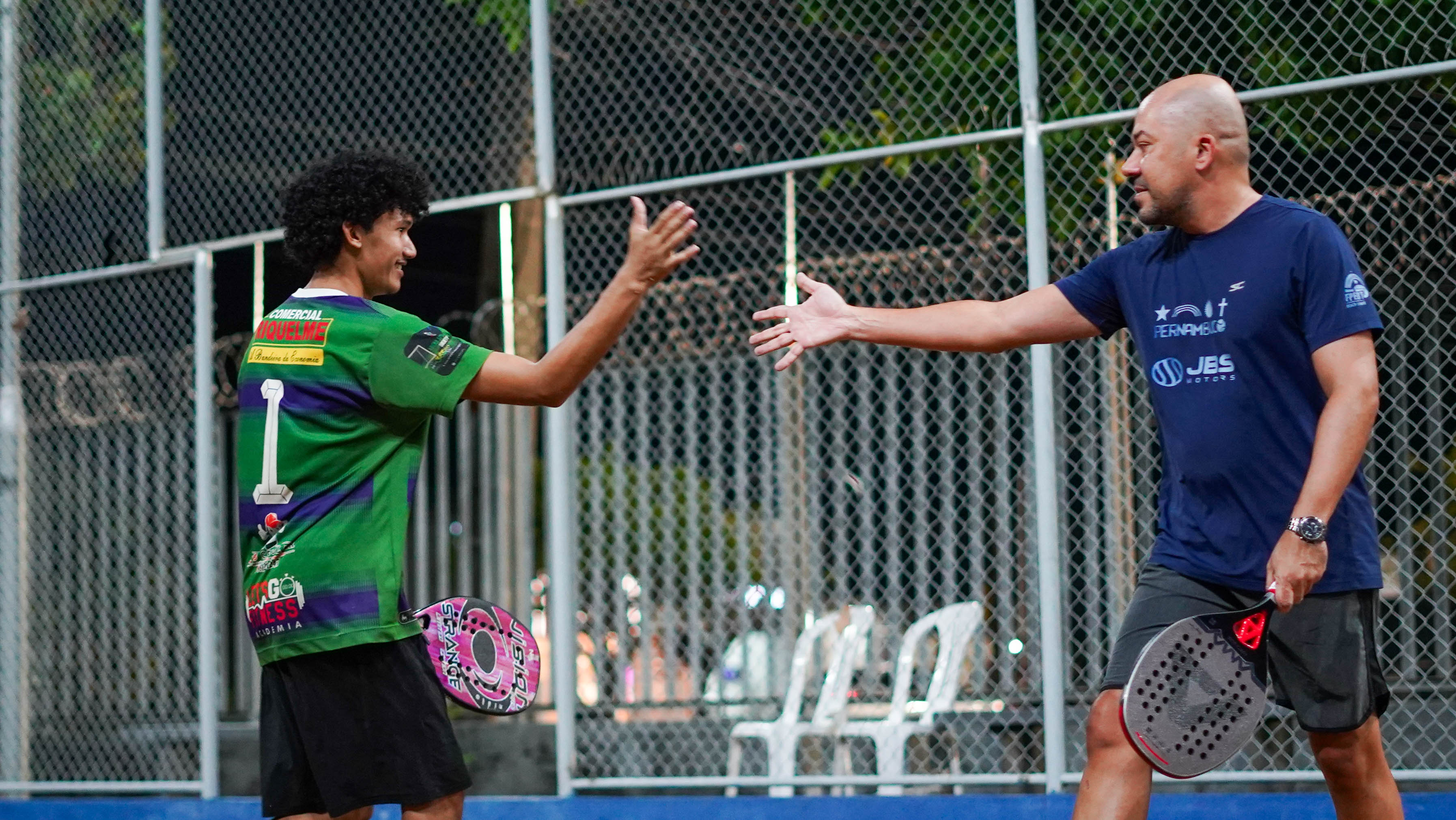 Imagem colorida mostra dois participantes do torneio dentro da quadra de areia. Os homens seguram raquetes e estendem as mãos um em direção ao outro em gesto de cumprimento. Ao fundo, aparecem a grade que cerca a quadra e cadeiras brancas. Imagem colorida mostra dois participantes do torneio dentro da quadra de areia. Os homens seguram raquetes e estendem as mãos um em direção ao outro em gesto de cumprimento. Ao fundo, aparecem a grade que cerca a quadra e cadeiras brancas.
