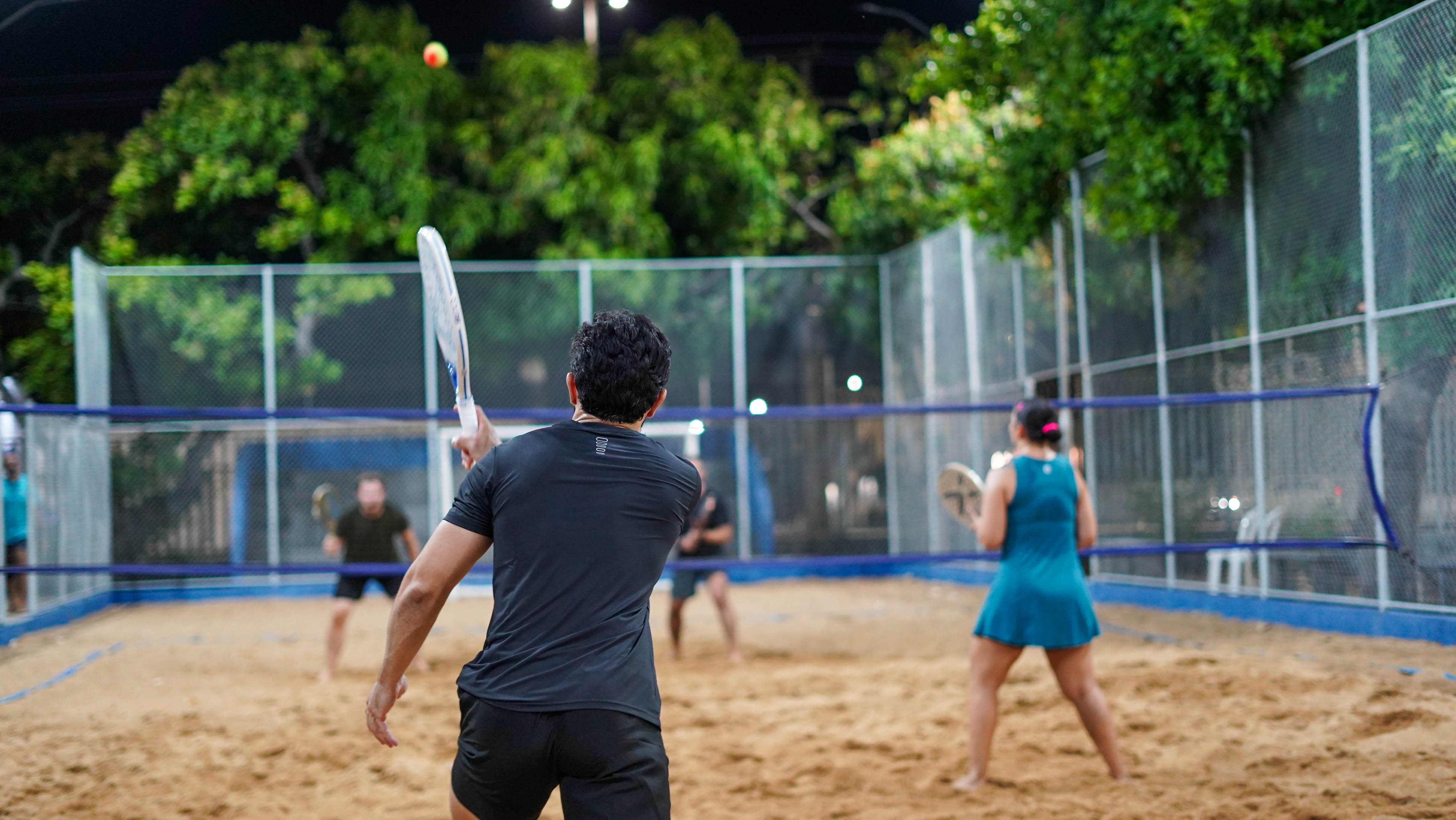 Imagem colorida mostra uma partida de beach tennis vista por trás de uma das duplas. Um homem de camiseta preta acompanha a bola com a raquete levantada, enquanto sua parceira, de roupa azul, está posicionada próxima à rede. Do outro lado, os adversários aguardam a jogada. Imagem colorida mostra uma partida de beach tennis vista por trás de uma das duplas. Um homem de camiseta preta acompanha a bola com a raquete levantada, enquanto sua parceira, de roupa azul, está posicionada próxima à rede. Do outro lado, os adversários aguardam a jogada.