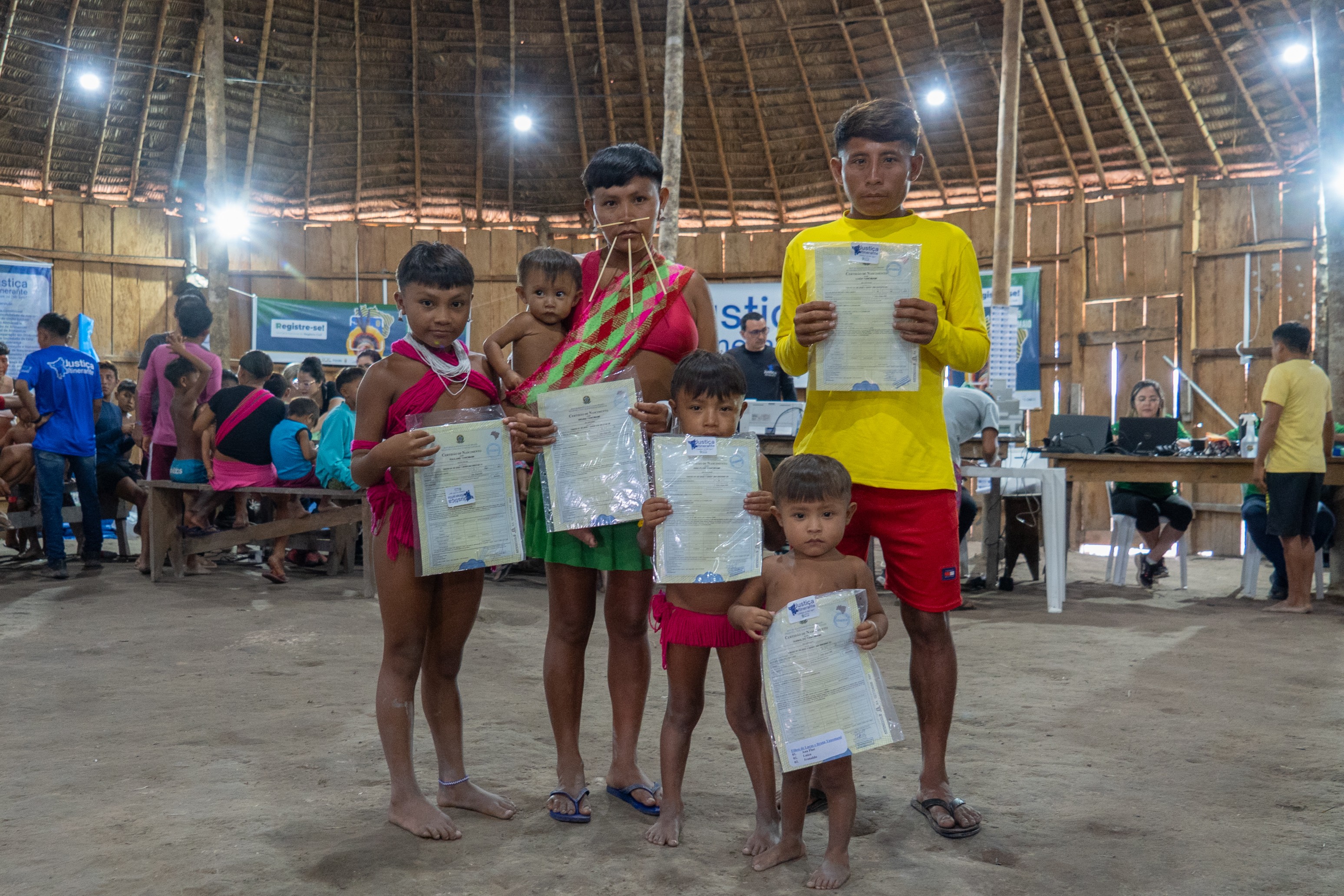 Imagem colorida mostra integrantes de uma família indígena dentro de uma maloca, segurando documentos emitidos durante uma ação da Justiça Itinerante. Duas mulheres, um homem e três crianças aparecem em primeiro plano, enquanto equipes prestam atendimento a outras pessoas ao fundo. O espaço possui estrutura de madeira e cobertura de palha, com mesas, computadores e banners da Justiça Itinerante.