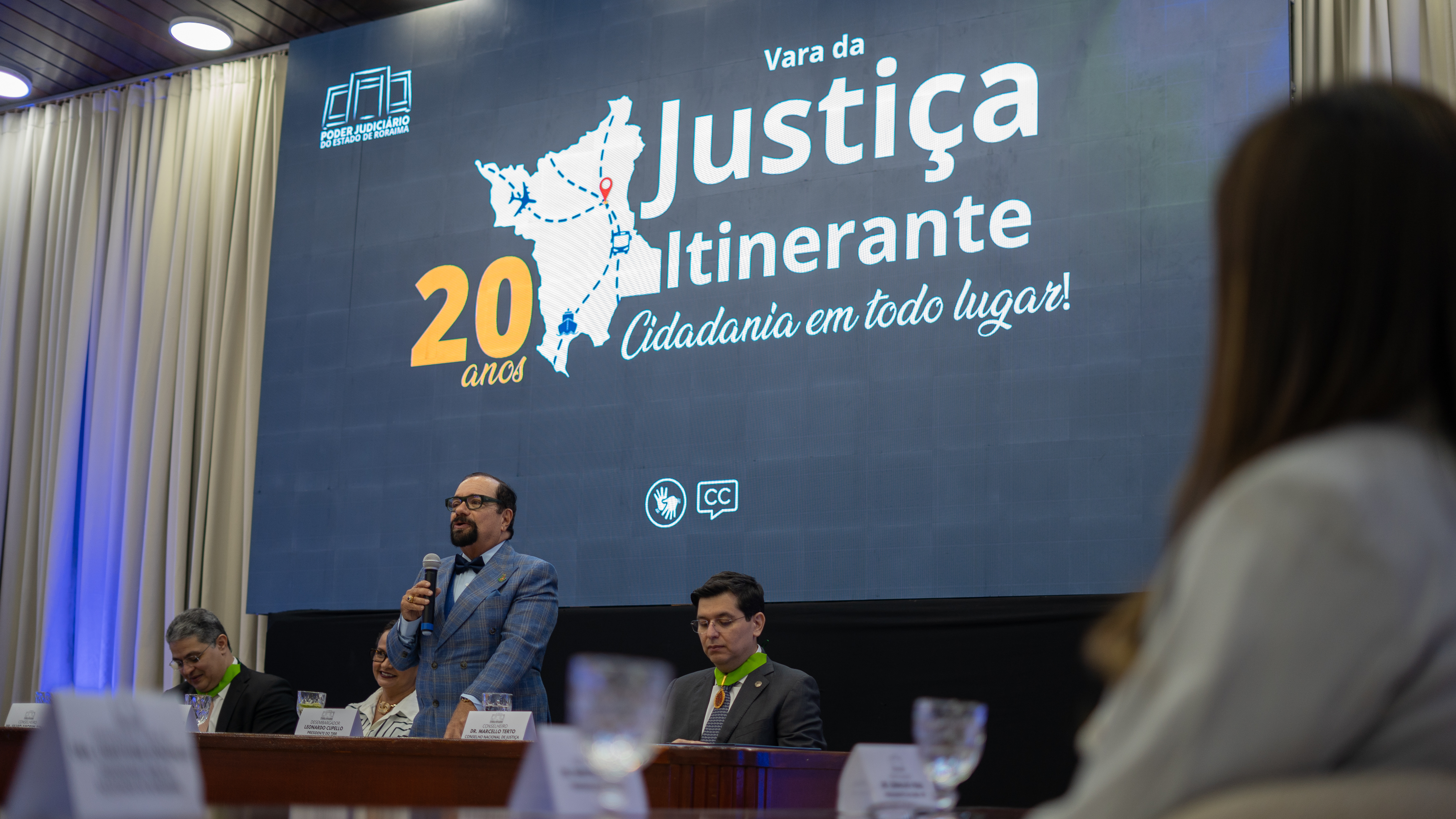 Imagem colorida mostra o presidente do TJRR, desembargador Leonardo Cupello, de pé atrás da mesa de autoridades, segurando um microfone durante discurso na solenidade. Ao fundo, o telão exibe a identidade visual dos 20 anos da Vara da Justiça Itinerante. Imagem colorida mostra o presidente do TJRR, desembargador Leonardo Cupello, de pé atrás da mesa de autoridades, segurando um microfone durante discurso na solenidade. Ao fundo, o telão exibe a identidade visual dos 20 anos da Vara da Justiça Itinerante.