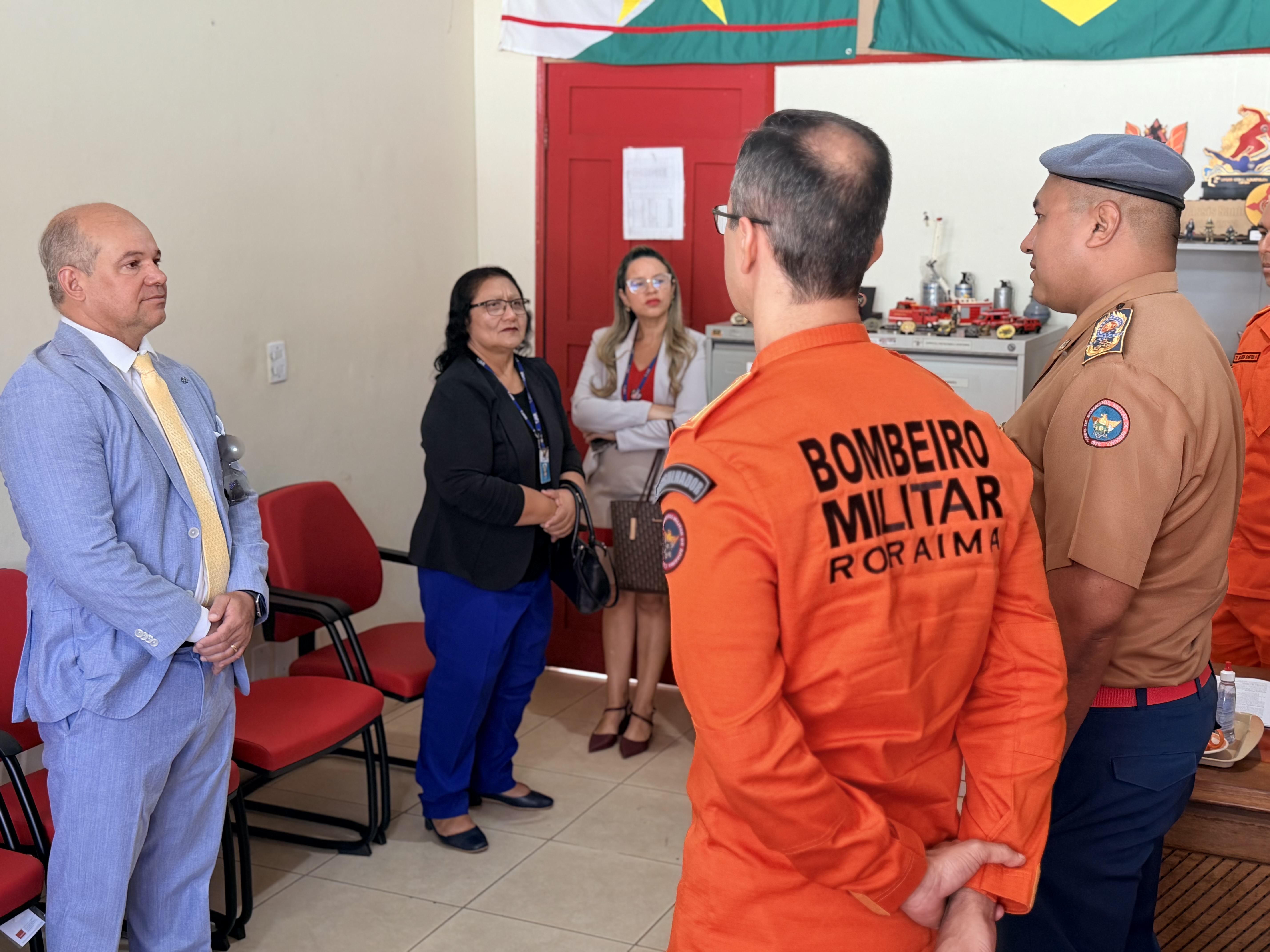: Em uma sala interna, o Dr. Eduardo de Carvalho e o Coronel Anderson Matos conversam com outras duas servidoras e um bombeiro de farda laranja. Na parede ao fundo, estão penduradas as bandeiras de Roraima e do Brasil.