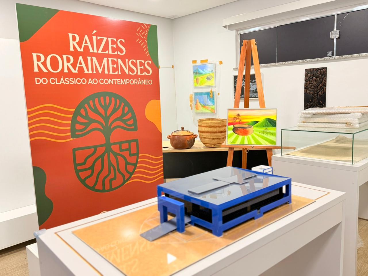 Imagem colorida mostra uma sala de exposição com painel vermelho escrito “Raízes Roraimenses – Do clássico ao contemporâneo”, ao lado de obras de arte, um cavalete com pintura de paisagem, peças de cerâmica, cestaria indígena e uma maquete arquitetônica azul sobre pedestal. Imagem colorida mostra uma sala de exposição com painel vermelho escrito “Raízes Roraimenses – Do clássico ao contemporâneo”, ao lado de obras de arte, um cavalete com pintura de paisagem, peças de cerâmica, cestaria indígena e uma maquete arquitetônica azul sobre pedestal.