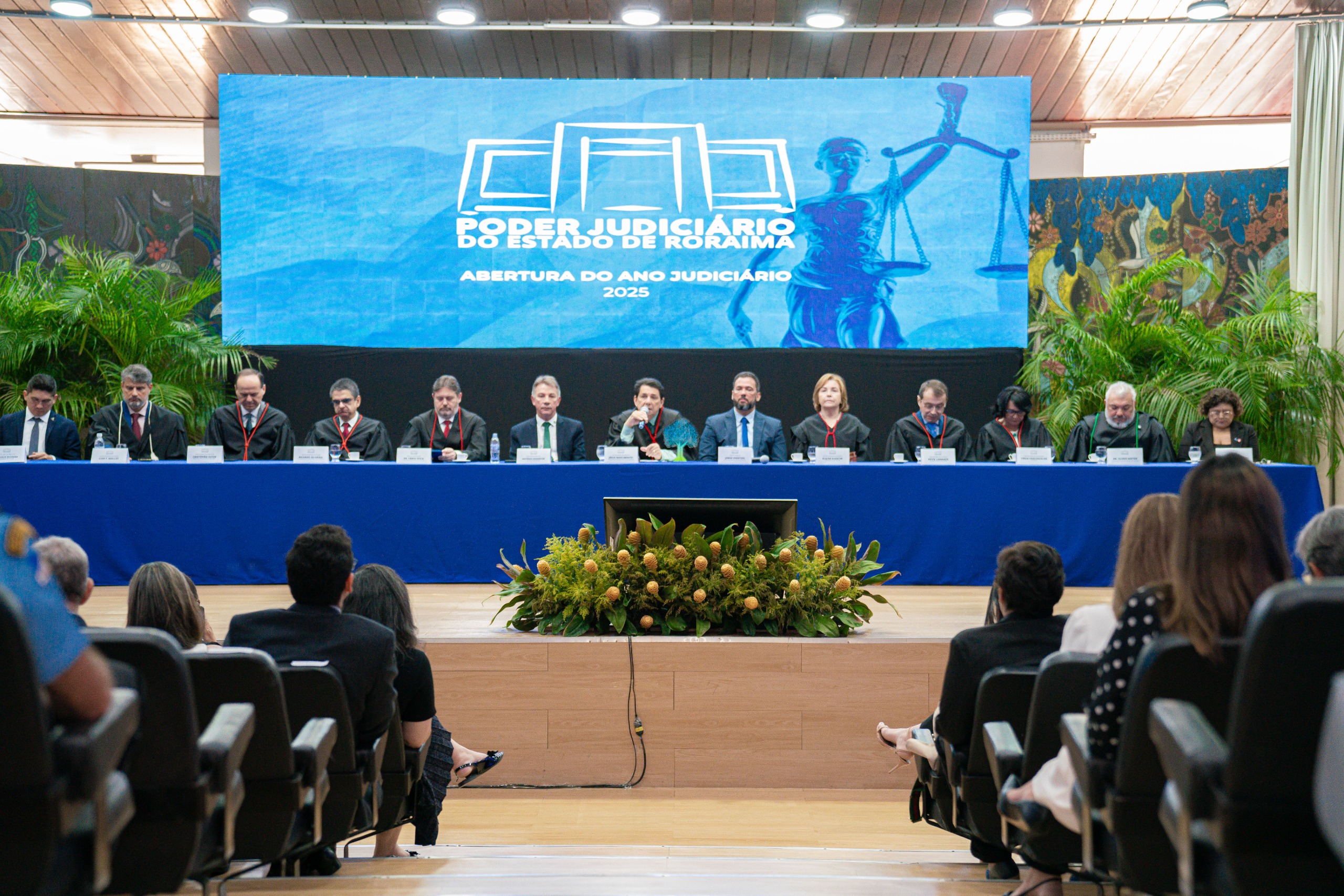 foto colorida mostra a solenidade de abertura do Ano Judiciário 2025 do Tribunal de Justiça de Roraima (TJRR). No palco, uma mesa com treze autoridades veste trajes formais e togas. Da esquerda para a direita: o presidente da Associação de Magistrados do Estado de Roraima, Marcelo Oliveira; o juiz convocado, Luiz Fernando Mallet; o diretor da Escola Judicial de Roraima, desembargador Cristóvão Súter; o vice-presidente do Tribunal de Justiça de Roraima, desembargador Ricardo Oliveira; o procurador-geral de Justiça do Ministério Público de Roraima, Fábio Stica; o governador do Estado de Roraima, Antonio Denarium; o presidente do Tribunal de Justiça do Estado de Roraima, desembargador Jésus Nascimento; o vice-presidente da Assembleia Legislativa do Estado de Roraima, deputado Jorge Everton; a presidente do TRE-Roraima, desembargadora Elaine Bianchi; o ouvidor-geral de Justiça, desembargador Erick Linhares; a corregedora do TRE, desembargadora Tânia Vasconcelos; o defensor público-geral do Estado de Roraima, Dr. Oleno Matos; e a vice-presidente da Ordem dos Advogados do Brasil – Seccional Roraima, Dra. Natália Leitão Costa. Ao fundo, um telão exibe o logotipo do TJRR e a inscrição "Abertura do Ano Judiciário 2025". foto colorida mostra a solenidade de abertura do Ano Judiciário 2025 do Tribunal de Justiça de Roraima (TJRR). No palco, uma mesa com treze autoridades veste trajes formais e togas. Da esquerda para a direita: o presidente da Associação de Magistrados do Estado de Roraima, Marcelo Oliveira; o juiz convocado, Luiz Fernando Mallet; o diretor da Escola Judicial de Roraima, desembargador Cristóvão Súter; o vice-presidente do Tribunal de Justiça de Roraima, desembargador Ricardo Oliveira; o procurador-geral de Justiça do Ministério Público de Roraima, Fábio Stica; o governador do Estado de Roraima, Antonio Denarium; o presidente do Tribunal de Justiça do Estado de Roraima, desembargador Jésus Nascimento; o vice-presidente da Assembleia Legislativa do Estado de Roraima, deputado Jorge Everton; a presidente do TRE-Roraima, desembargadora Elaine Bianchi; o ouvidor-geral de Justiça, desembargador Erick Linhares; a corregedora do TRE, desembargadora Tânia Vasconcelos; o defensor público-geral do Estado de Roraima, Dr. Oleno Matos; e a vice-presidente da Ordem dos Advogados do Brasil – Seccional Roraima, Dra. Natália Leitão Costa. Ao fundo, um telão exibe o logotipo do TJRR e a inscrição "Abertura do Ano Judiciário 2025".