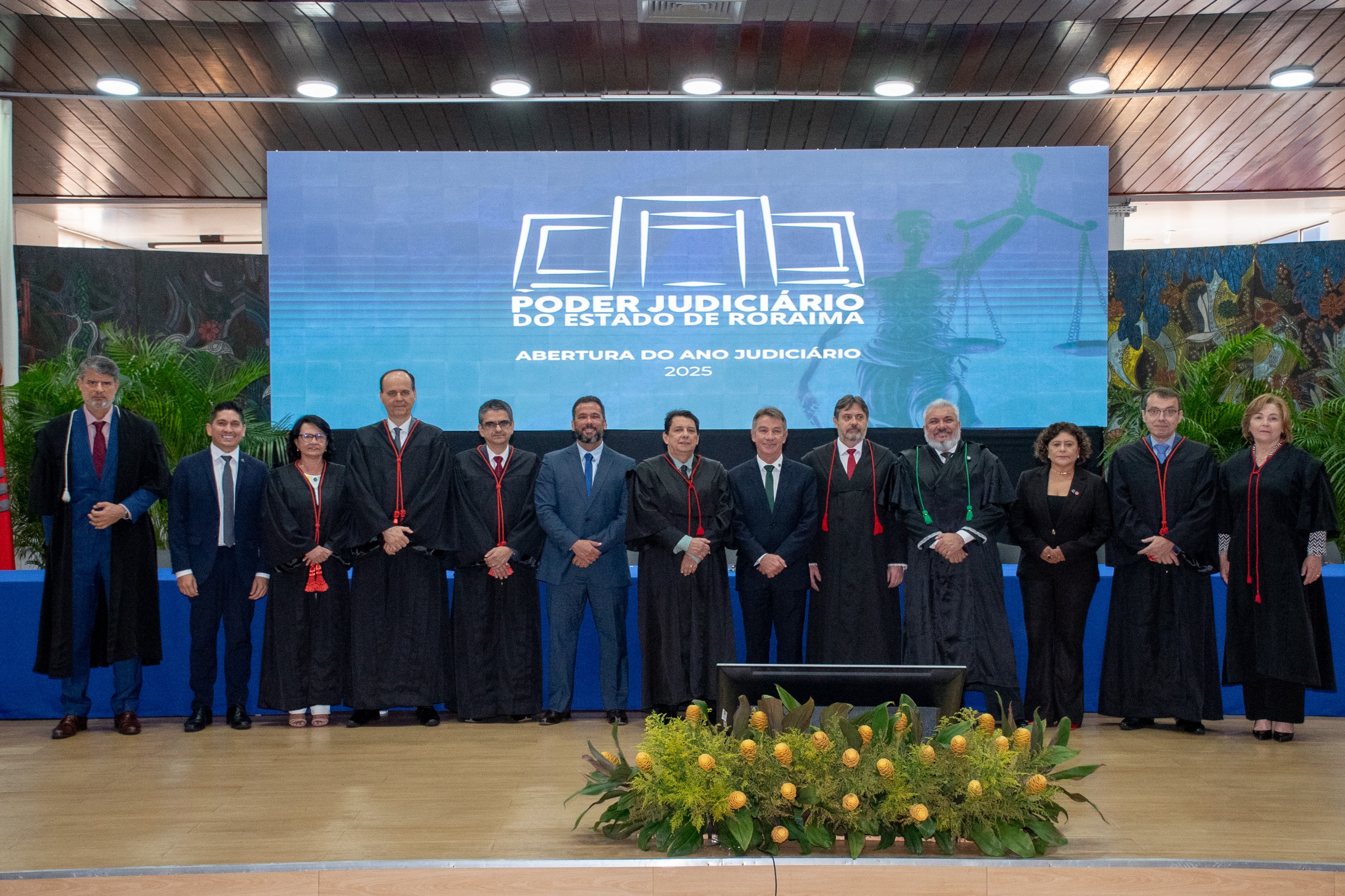 foto colorida mostra treze autoridades posando em pé para uma foto oficial durante a abertura do Ano Judiciário 2025 do Tribunal de Justiça de Roraima. Da esquerda para a dirita: o juiz convocado, Luiz Fernando Mallet; o presidente da Associação de Magistrados do Estado de Roraima, Marcelo Oliveira; a corregedora do TRE, desembargadora Tânia Vasconcelos; o diretor da Escola Judicial de Roraima, desembargador Cristóvão Súter; o vice-presidente do Tribunal de Justiça de Roraima, desembargador Ricardo Oliveira; o vice-presidente da Assembleia Legislativa do Estado de Roraima, deputado Jorge Everton; o vice-presidente da Assembleia Legislativa do Estado de Roraima, deputado Jorge Everton; o presidente do Tribunal de Justiça do Estado de Roraima, desembargador Jésus Nascimento; o governador do Estado de Roraima, Antônio Denarium; o procurador-geral de Justiça do Ministério Público de Roraima, Fábio Stica; o defensor público-geral do Estado de Roraima, Dr. Oleno Matos; a vice-presidente da Ordem dos Advogados do Brasil – Seccional Roraima, Dra. Natália Leitão Costa; o ouvidor-geral de Justiça, desembargador Erick Linhares; e a presidente do TRE-Roraima, desembargadora Elaine Bianchi. Eles estão no palco, vestidos com trajes formais e togas, à frente de um telão que exibe o logotipo do TJRR e a inscrição "Abertura do Ano Judiciário 2025". foto colorida mostra treze autoridades posando em pé para uma foto oficial durante a abertura do Ano Judiciário 2025 do Tribunal de Justiça de Roraima. Da esquerda para a dirita: o juiz convocado, Luiz Fernando Mallet; o presidente da Associação de Magistrados do Estado de Roraima, Marcelo Oliveira; a corregedora do TRE, desembargadora Tânia Vasconcelos; o diretor da Escola Judicial de Roraima, desembargador Cristóvão Súter; o vice-presidente do Tribunal de Justiça de Roraima, desembargador Ricardo Oliveira; o vice-presidente da Assembleia Legislativa do Estado de Roraima, deputado Jorge Everton; o vice-presidente da Assembleia Legislativa do Estado de Roraima, deputado Jorge Everton; o presidente do Tribunal de Justiça do Estado de Roraima, desembargador Jésus Nascimento; o governador do Estado de Roraima, Antônio Denarium; o procurador-geral de Justiça do Ministério Público de Roraima, Fábio Stica; o defensor público-geral do Estado de Roraima, Dr. Oleno Matos; a vice-presidente da Ordem dos Advogados do Brasil – Seccional Roraima, Dra. Natália Leitão Costa; o ouvidor-geral de Justiça, desembargador Erick Linhares; e a presidente do TRE-Roraima, desembargadora Elaine Bianchi. Eles estão no palco, vestidos com trajes formais e togas, à frente de um telão que exibe o logotipo do TJRR e a inscrição "Abertura do Ano Judiciário 2025".