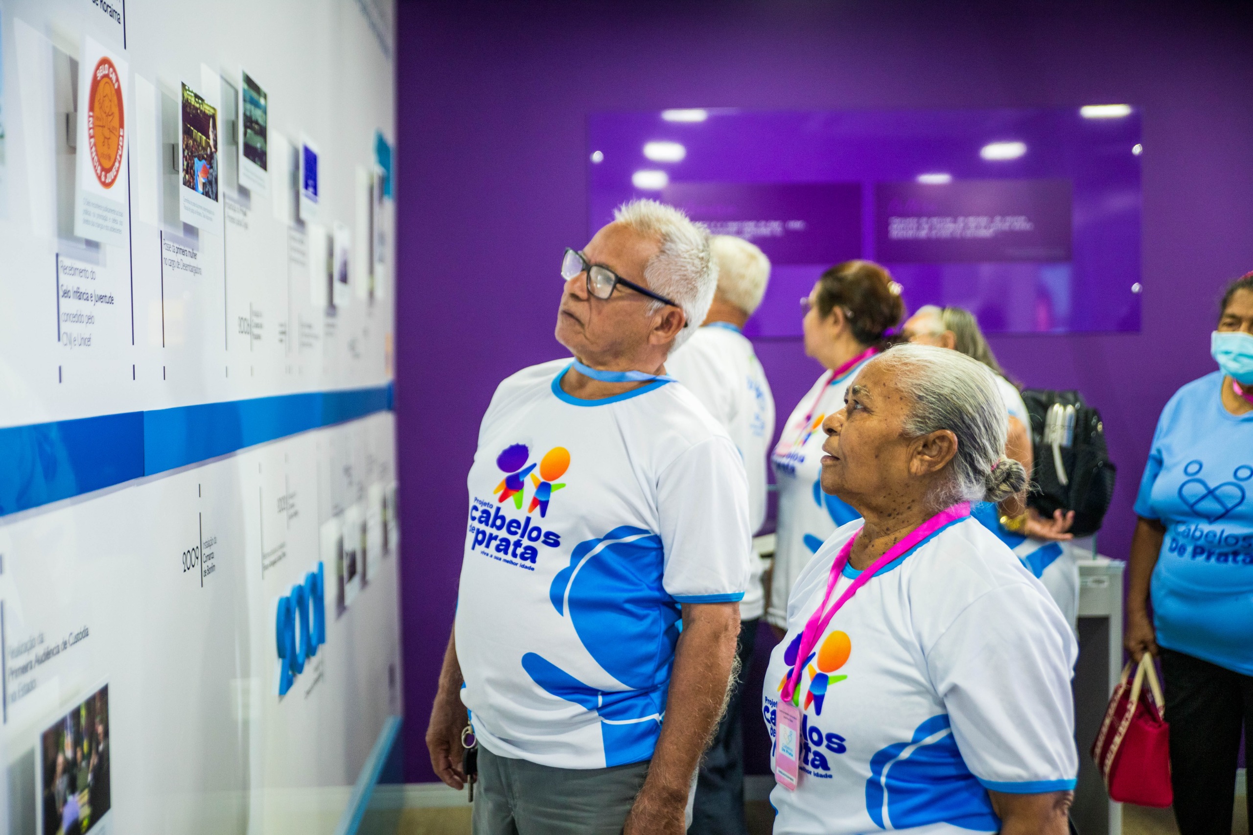 imagem colorida contém um casal de idosos observando a linha do tempo destacada na parede do CMC/TJRR, durante visita do grupo cabelos de prata. imagem colorida contém um casal de idosos observando a linha do tempo destacada na parede do CMC/TJRR, durante visita do grupo cabelos de prata.