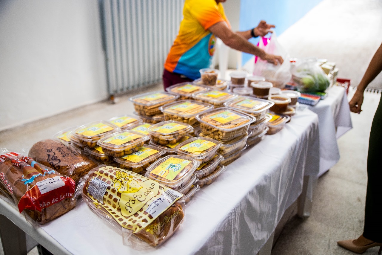 A imagem retrata uma mesa com diversos alimentos embalados, como pães, biscoitos e potes com grãos ou preparações, sugerindo uma ação de distribuição alimentar ou venda comunitária.