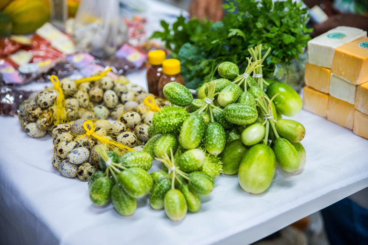 A imagem mostra uma mesa de feira com alimentos frescos e produtos locais em Boa Vista (RR), incluindo cucamelons (mini pepinos melancia), ovos de codorna, ervas frescas, potes com mel ou conservas e queijos embalados.