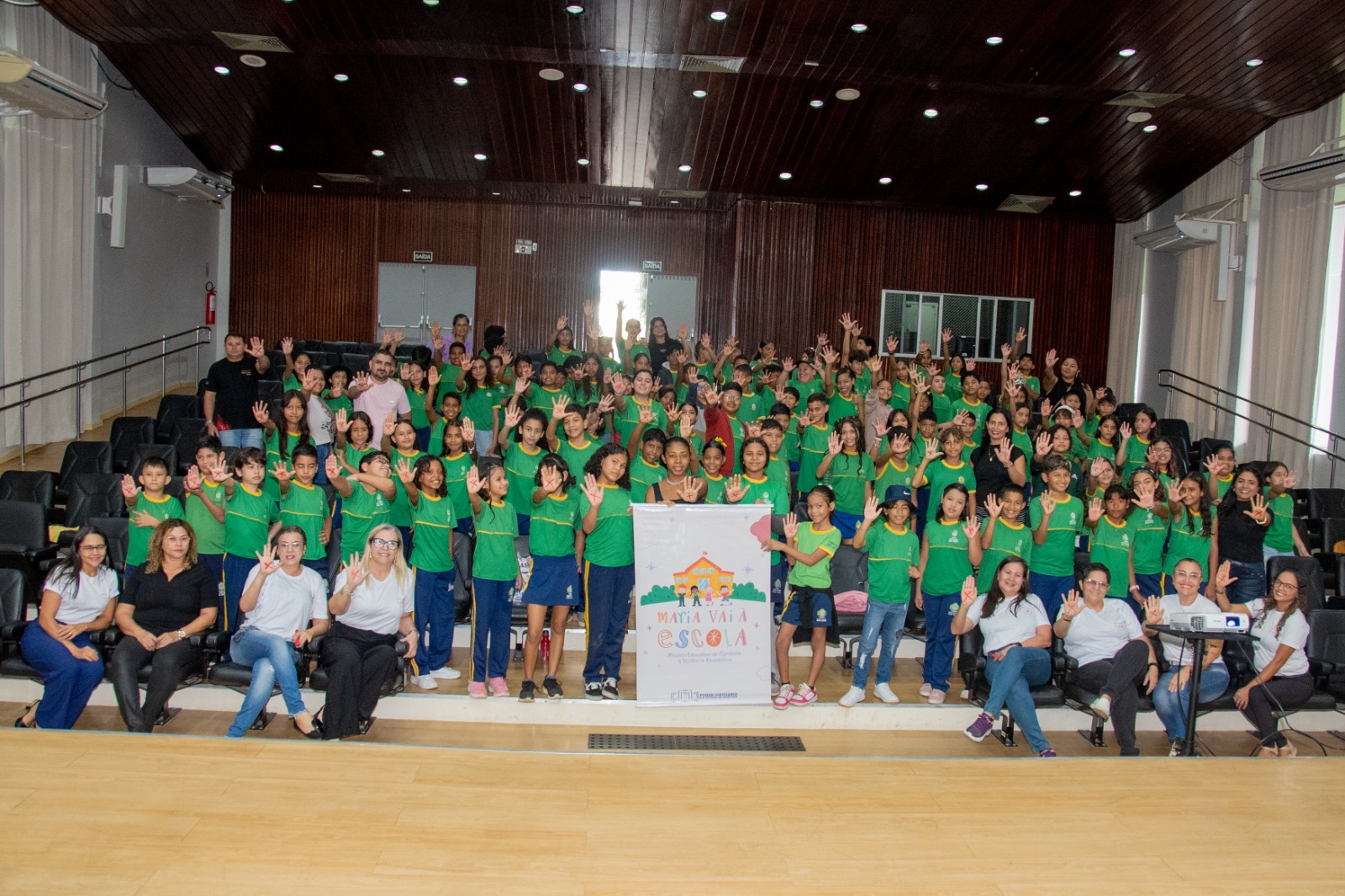 Foto colorida mostra um auditório com crianças uniformizadas e adultos. A maioria está de pé, estendendo a mão, como o simbolo usado pelas mulheres que sofrem agressão e buscam ajuda. Foto colorida mostra um auditório com crianças uniformizadas e adultos. A maioria está de pé, estendendo a mão, como o simbolo usado pelas mulheres que sofrem agressão e buscam ajuda.