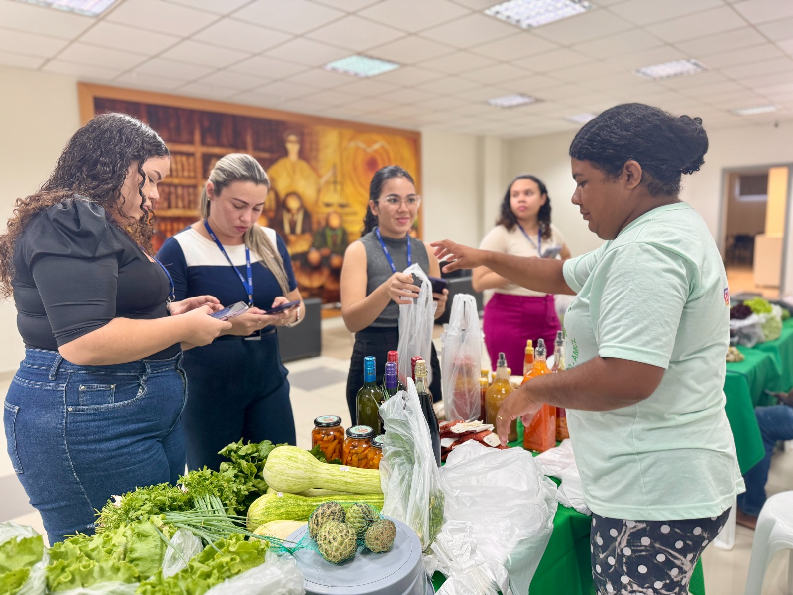 Foto colorida mostrando quatro servidoras do TJRR comprando legumes com duas mulheres das associações parceiras da Feira de Orgânicos. Abaixo na foto uma mesa cheia de produtos. Foto colorida mostrando quatro servidoras do TJRR comprando legumes com duas mulheres das associações parceiras da Feira de Orgânicos. Abaixo na foto uma mesa cheia de produtos.