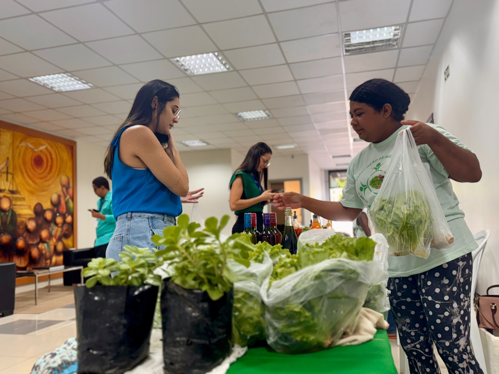Foto colorida mostrando duas servidoras do TJRR comprando legumes com duas mulheres das associações parceiras da Feira de Orgânicos. Abaixo na foto uma mesa cheia de produtos Foto colorida mostrando duas servidoras do TJRR comprando legumes com duas mulheres das associações parceiras da Feira de Orgânicos. Abaixo na foto uma mesa cheia de produtos