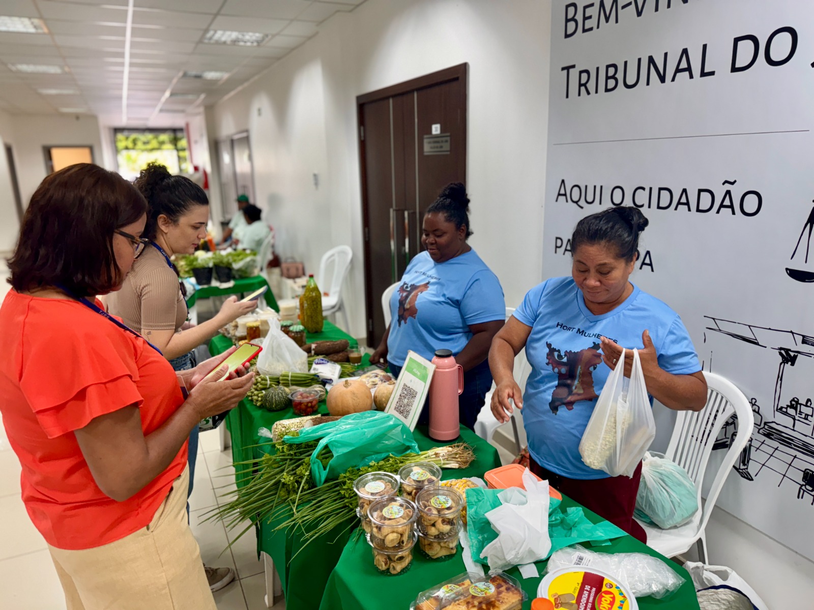 Foto colorida mostrando duas servidoras do TJRR comprando legumes com duas mulheres das associações parceiras da Feira de Orgânicos. Abaixo na foto uma mesa cheia de produtos. Foto colorida mostrando duas servidoras do TJRR comprando legumes com duas mulheres das associações parceiras da Feira de Orgânicos. Abaixo na foto uma mesa cheia de produtos.