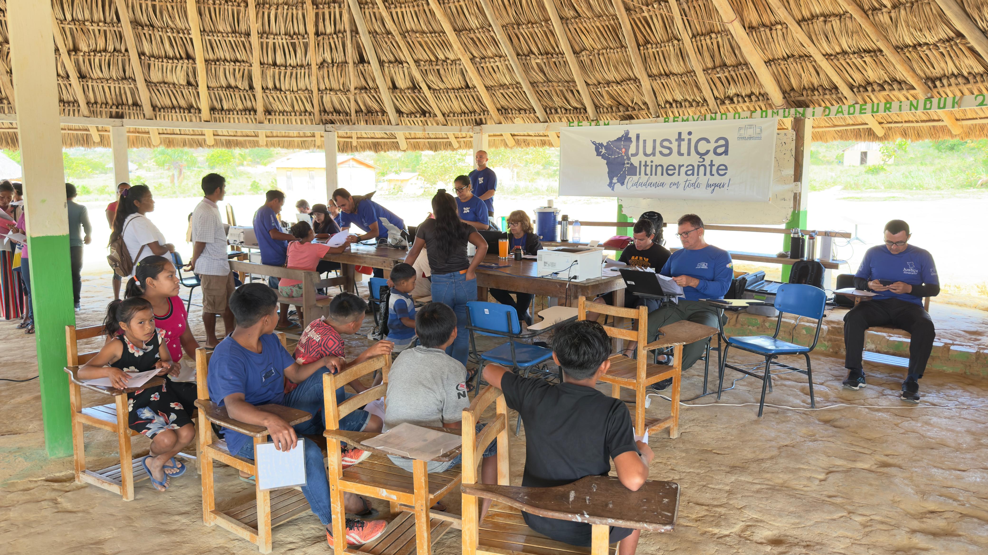 Imagem colorida registrando o atendimento da Vara da Justiça Itinerante do TJRR à comunidade indígena Urunduk, em Uiramutã, sob um tapiri. Na cena, os servidores realizam atendimentos enquanto os membros da comunidade aguardam, organizados em fila ou sentados em cadeiras.
