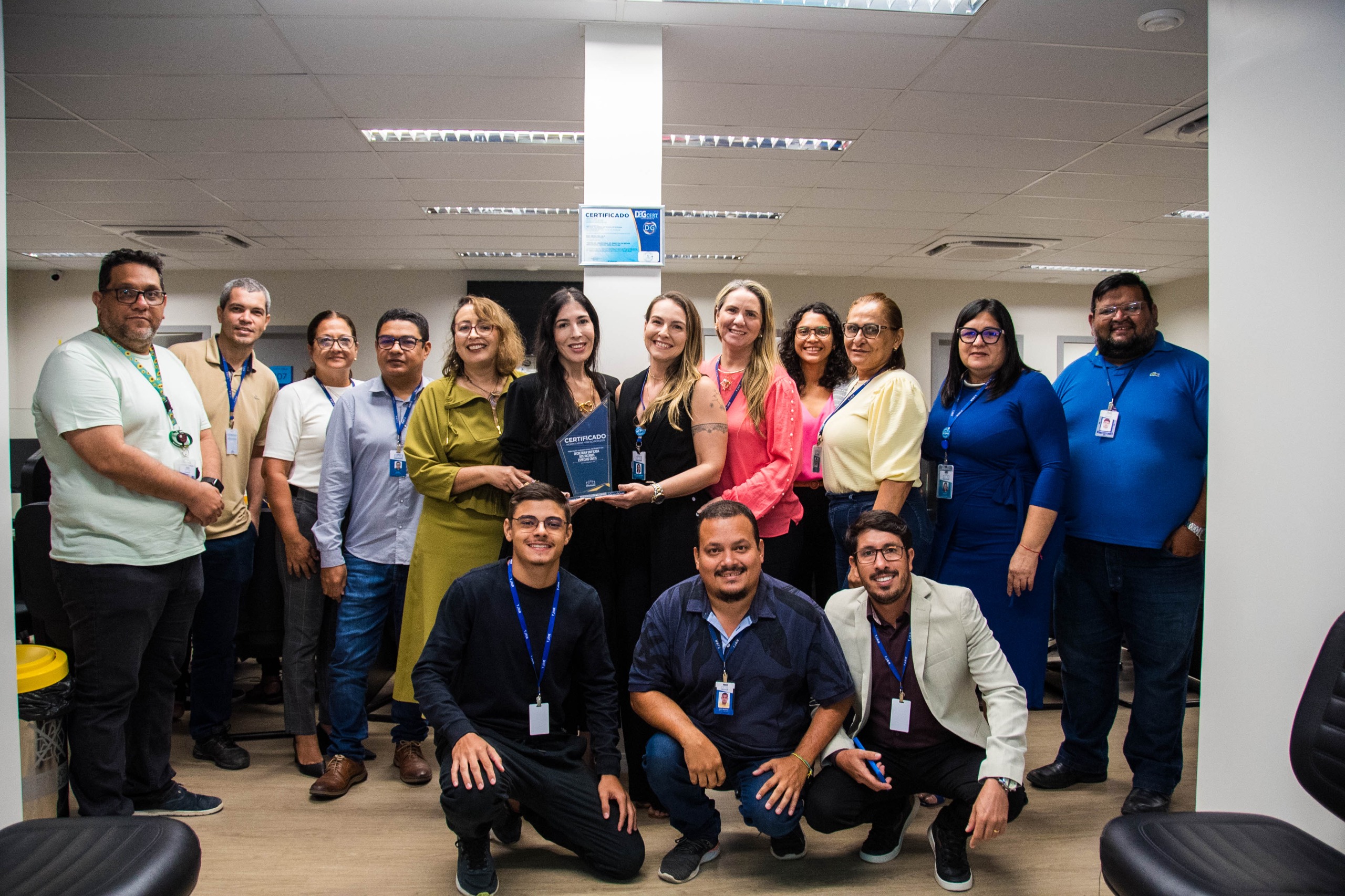 Imagem colorida contém a juíza Suelen Márcia Silva Alves, titular do 1.º Juizado Especializado de Violência Doméstica e Familiar contra a Mulher, segurando placa de premiação da certificação do ISO 9001, e posando para fotografia ao lado da Juíza Auxiliar da Presidência do TJRR, Lana Leitão, da Subsecretária de Processos e Gestão da Qualidade, Erika Horta e mais sete servidores do TJRR.