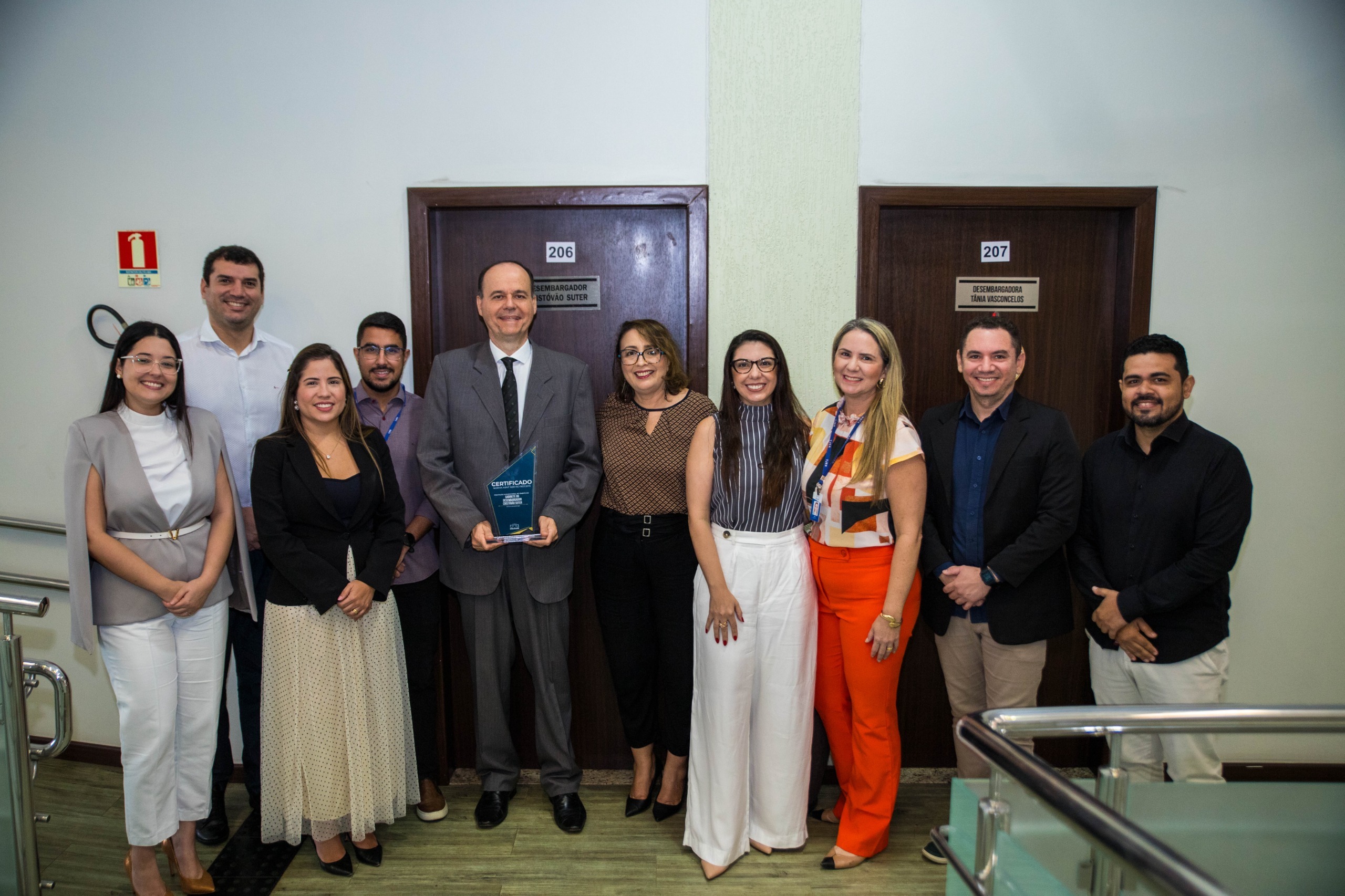 Imagem colorida contém Desembargador Cristóvão Suter,segurando placa de premiação da certificação do ISO 9001, e posando para fotografia ao lado da Juíza Auxiliar da Presidência do TJRR, Lana Leitão, da Subsecretária de Processos e Gestão da Qualidade, Erika Horta e mais sete servidores do TJRR, em frente a entrada gabinete do Desembargador Cristóvão, localizado no Palácio da Justiça.