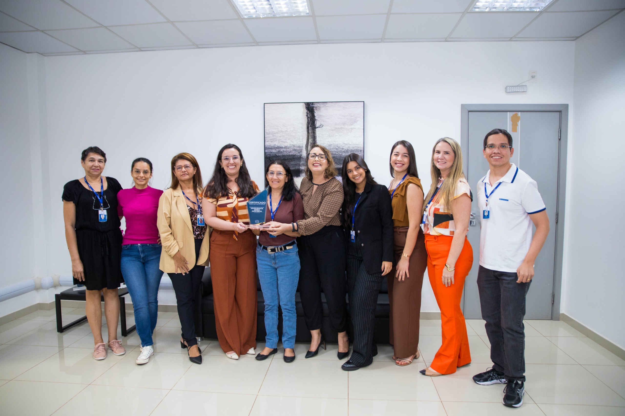 Imagem colorida contém a juíza titular do 3º Juizado Especial Cível, Bruna Guimarães Bezerra Fialho,segurando placa de premiação da certificação do ISO 9001, e posando para fotografia ao lado da Juíza Auxiliar da Presidência do TJRR, Lana Leitão, da Subsecretária de Processos e Gestão da Qualidade, Erika Horta e mais 12 servidores do TJRR.
