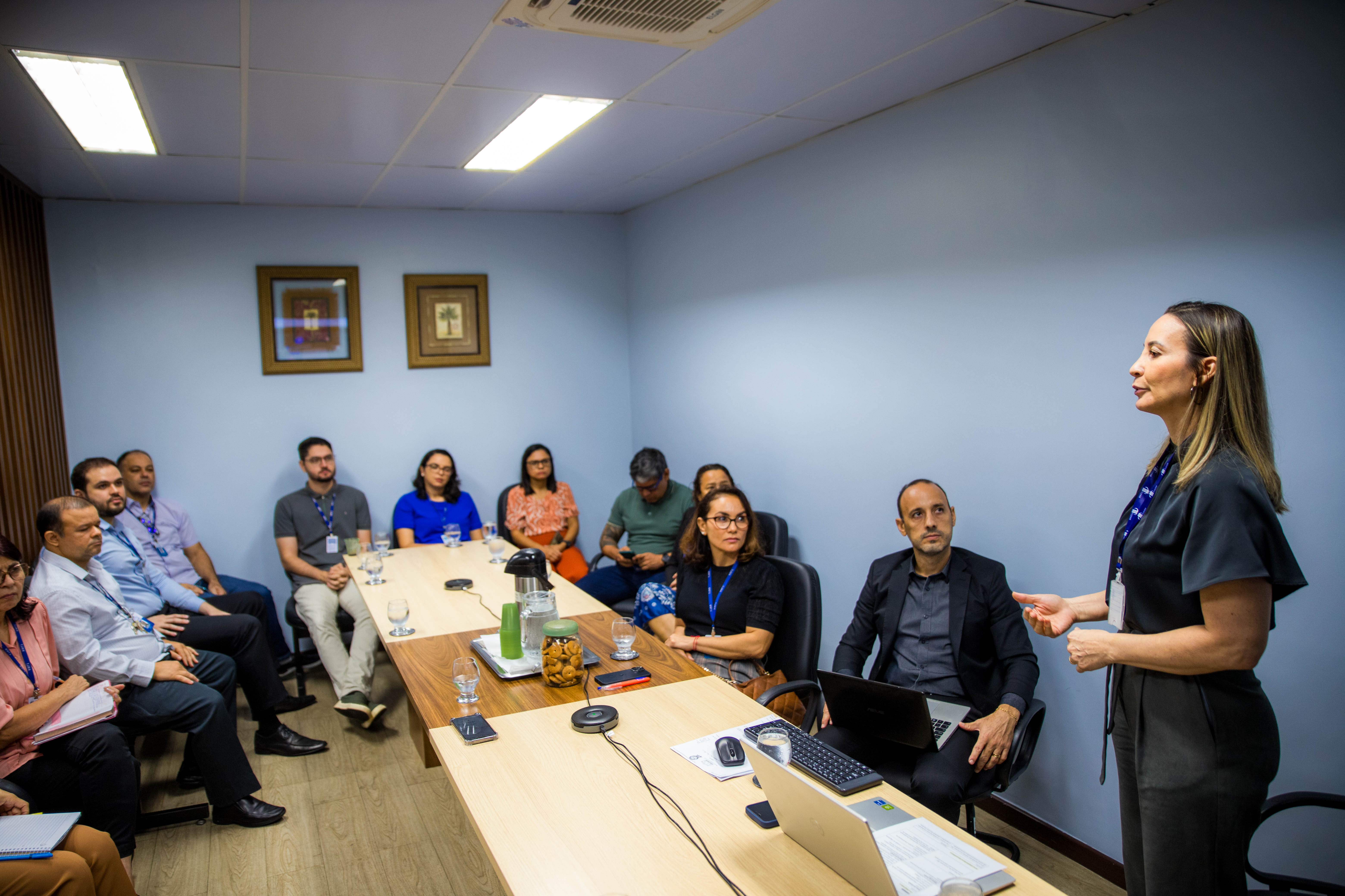 Imagem colorida contém uma mulher em pé na ponta de uma mesa, apresentando slide para servidores que se encontram presencial e outros em formato online por meio do meet. Ao redor da imagem encontram-se cinco servidores durante reunião da implementação do Sei Julgar, que ocorreu na sala de Togas do Tribunal Pleno do TJRR.