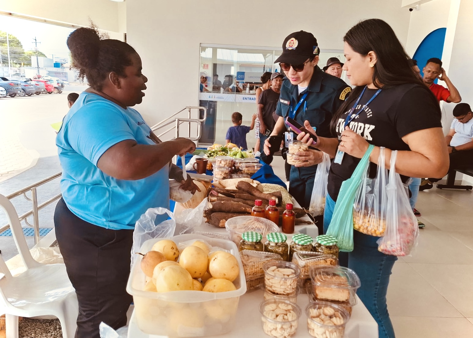 Foto colorida em plano aberto de uma feirante, mulher com uma blusa azul vibrante, atendendo duas clientes em uma mesa repleta de produtos naturais, como frutas, legumes, compotas e castanhas. As clientes, uma vestindo um uniforme escuro e a outra com uma camiseta preta, mostram interesse pelos produtos expostos. Foto colorida em plano aberto de uma feirante, mulher com uma blusa azul vibrante, atendendo duas clientes em uma mesa repleta de produtos naturais, como frutas, legumes, compotas e castanhas. As clientes, uma vestindo um uniforme escuro e a outra com uma camiseta preta, mostram interesse pelos produtos expostos.