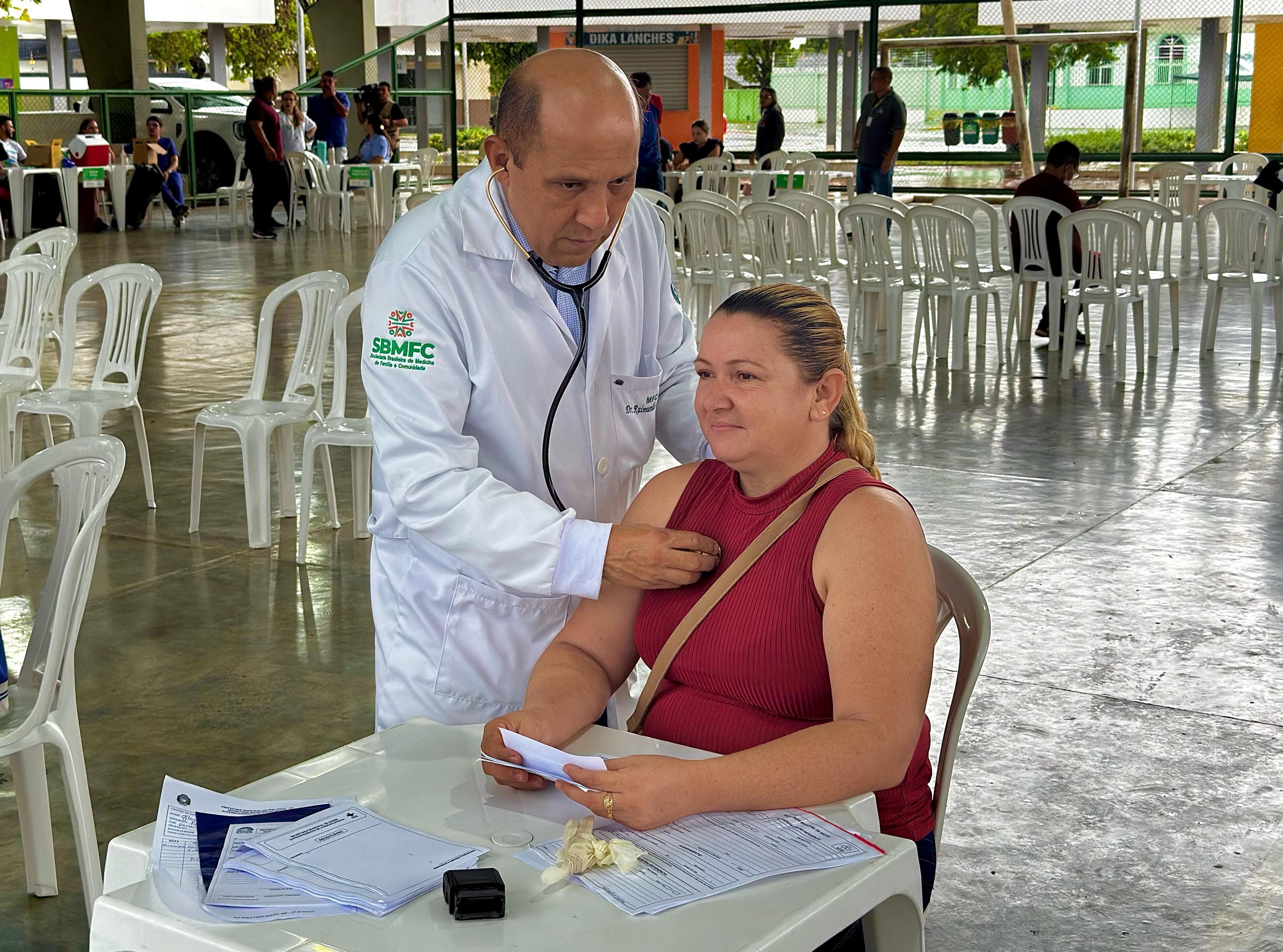 Foto colorida mostrando um médico, examinando uma paciente sentada. O médico está usando um estetoscópio para auscultar o peito da mulher. A paciente está vestindo uma blusa vermelha e tem uma bolsa a tiracolo. Ela parece estar segurando alguns papéis em suas mãos. Foto colorida mostrando um médico, examinando uma paciente sentada. O médico está usando um estetoscópio para auscultar o peito da mulher. A paciente está vestindo uma blusa vermelha e tem uma bolsa a tiracolo. Ela parece estar segurando alguns papéis em suas mãos.