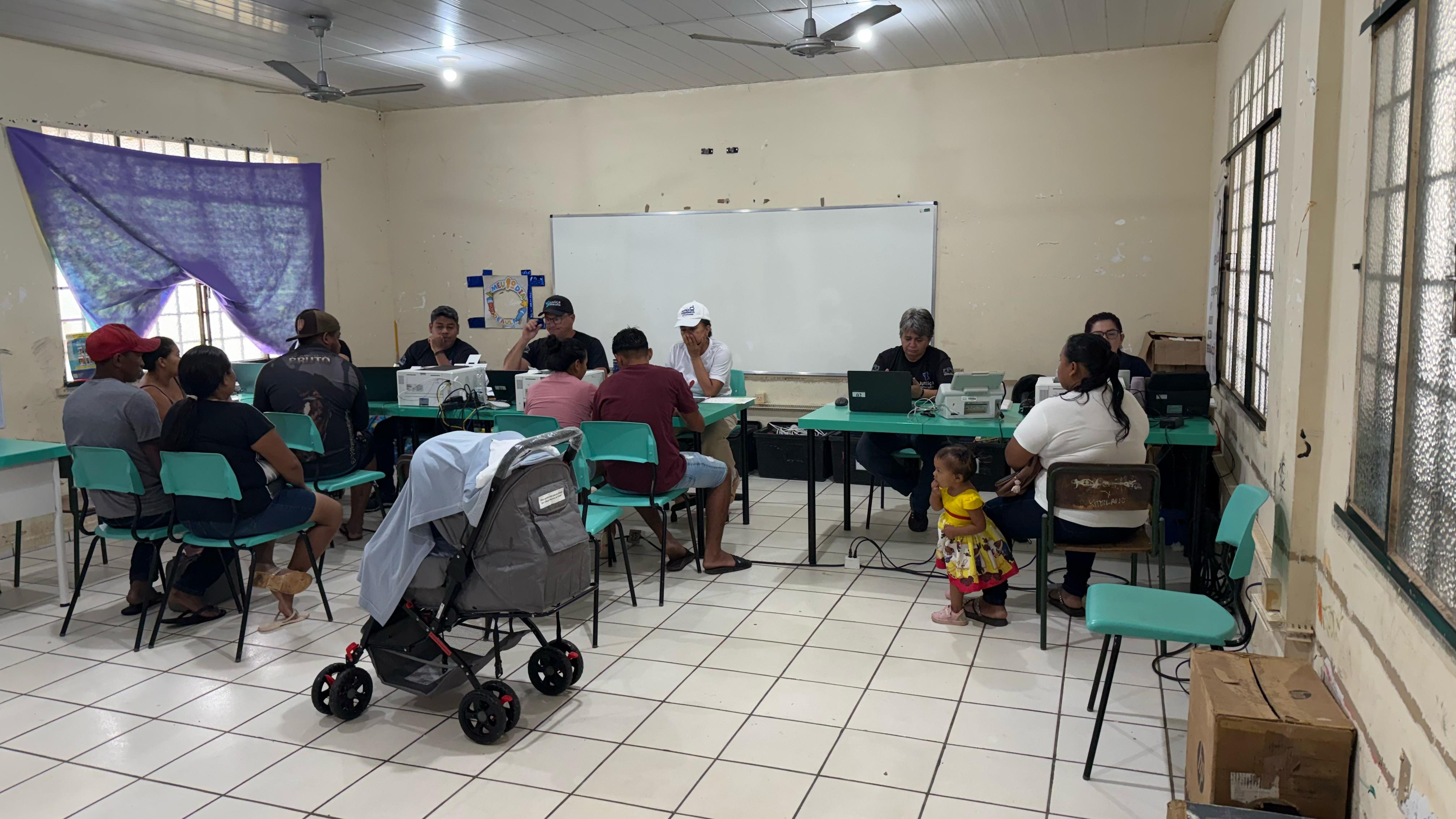 Imagem colorida mostra cerca de sete cidadãos sendo atendidos pela Vara da Justiça Itinerante do TJRR. É possível ver seis servidores sentados de frente para a população, realizando os atendimentos em uma sala de aula. Uma das cidadãs está com um bebê ao lado, em um carrinho, e outra com sua filha ao lado enquanto aguarda atendimento. Imagem colorida mostra cerca de sete cidadãos sendo atendidos pela Vara da Justiça Itinerante do TJRR. É possível ver seis servidores sentados de frente para a população, realizando os atendimentos em uma sala de aula. Uma das cidadãs está com um bebê ao lado, em um carrinho, e outra com sua filha ao lado enquanto aguarda atendimento.