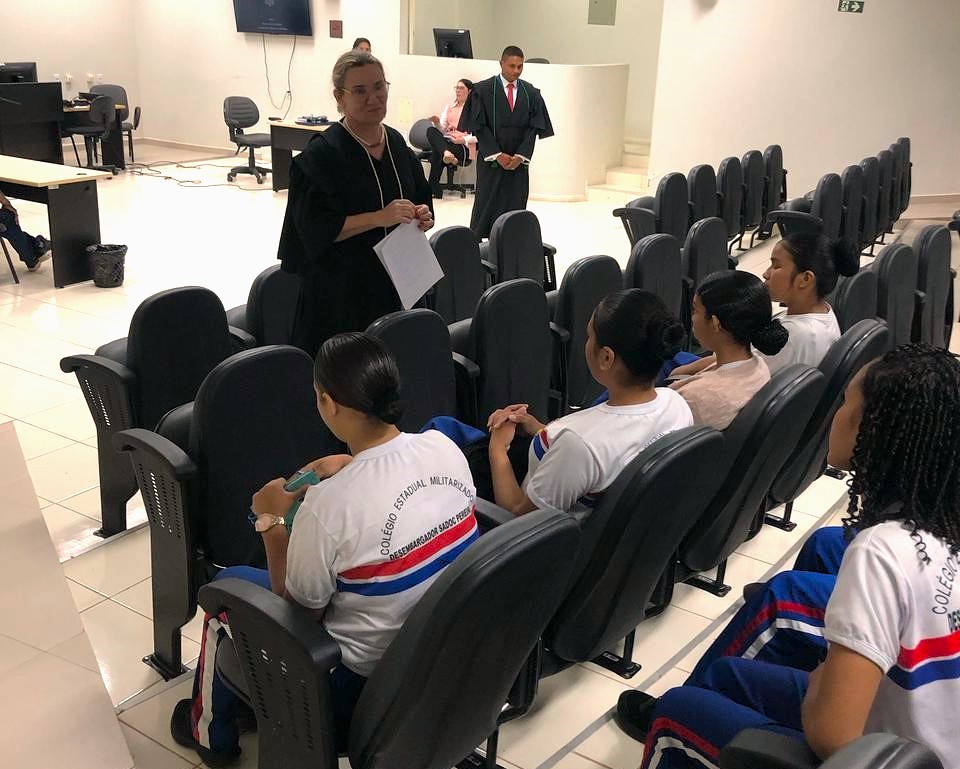 A imagem mostra o auditorio da comarca de Alto Alegre. No centro da imagem está a juíza titular da unidade, Sissi Marlene, vestida com uma toga preta e branca enquanto estudantes estão sentado à frente nas poltronas do auditório A imagem mostra o auditorio da comarca de Alto Alegre. No centro da imagem está a juíza titular da unidade, Sissi Marlene, vestida com uma toga preta e branca enquanto estudantes estão sentado à frente nas poltronas do auditório