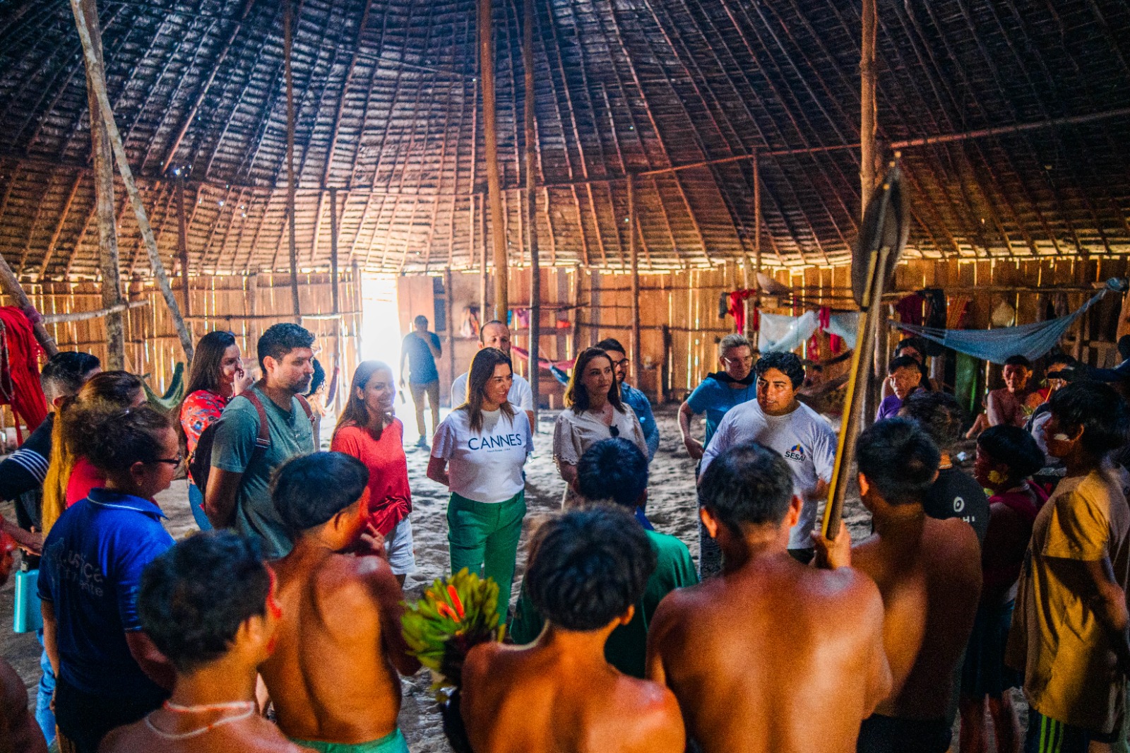 A imagem mostra um grupo diversificado de pessoas reunidas dentro de uma estrutura indígena tradicional. No centro da imagem, há um grupo de pessoas conversando. 