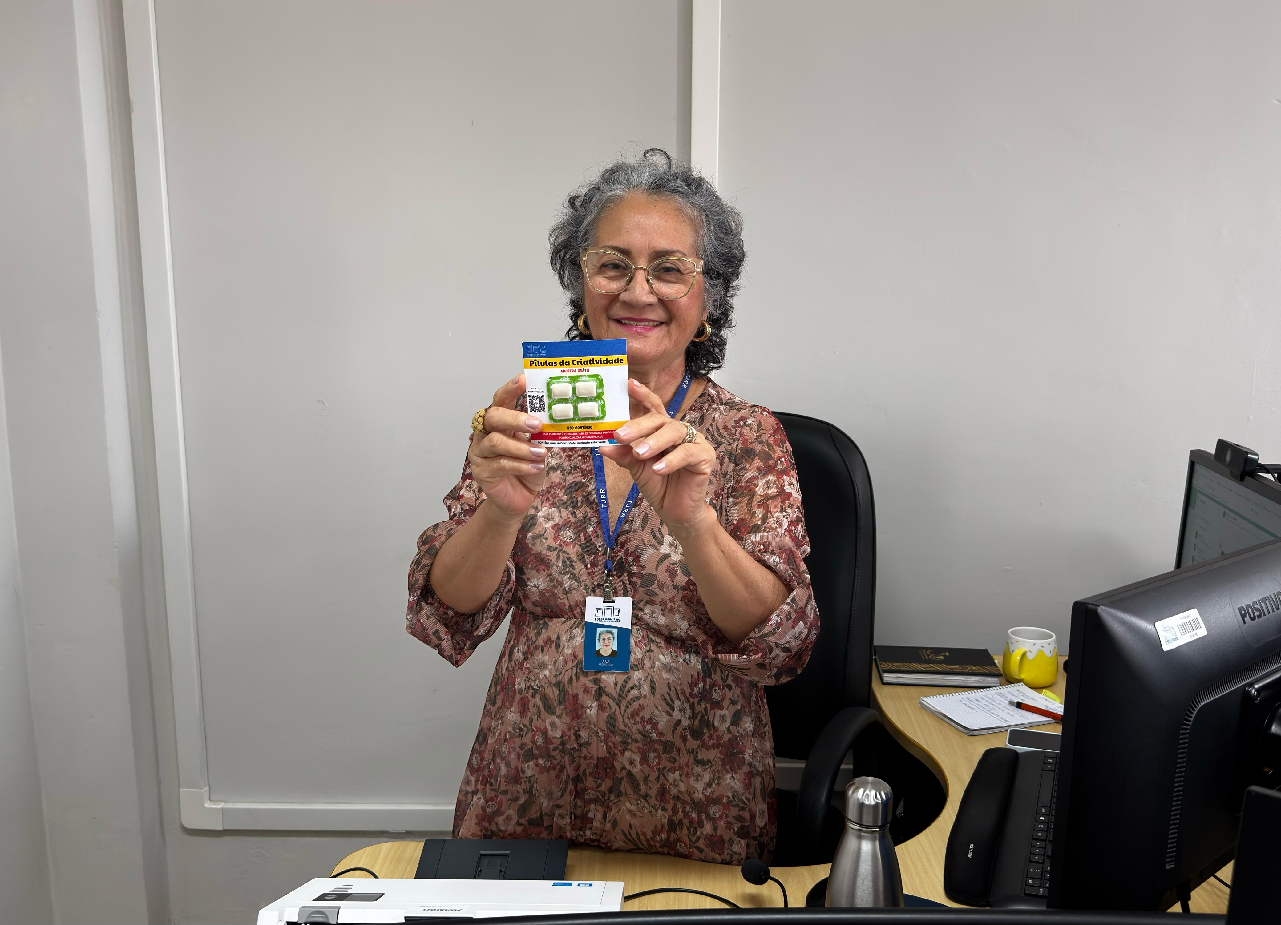 A imagem mostra uma mulher de cabelos grisalhos e óculos, sorrindo e segurando uma embalagem do produto "Pílulas da Criatividade" com ambas as mãos, em frente ao seu corpo. Ela está sentada em uma cadeira de escritório, atrás de uma mesa com um computador, um telefone preto, uma garrafa de água de metal e outros itens de escritório parcialmente visíveis. A imagem mostra uma mulher de cabelos grisalhos e óculos, sorrindo e segurando uma embalagem do produto "Pílulas da Criatividade" com ambas as mãos, em frente ao seu corpo. Ela está sentada em uma cadeira de escritório, atrás de uma mesa com um computador, um telefone preto, uma garrafa de água de metal e outros itens de escritório parcialmente visíveis.