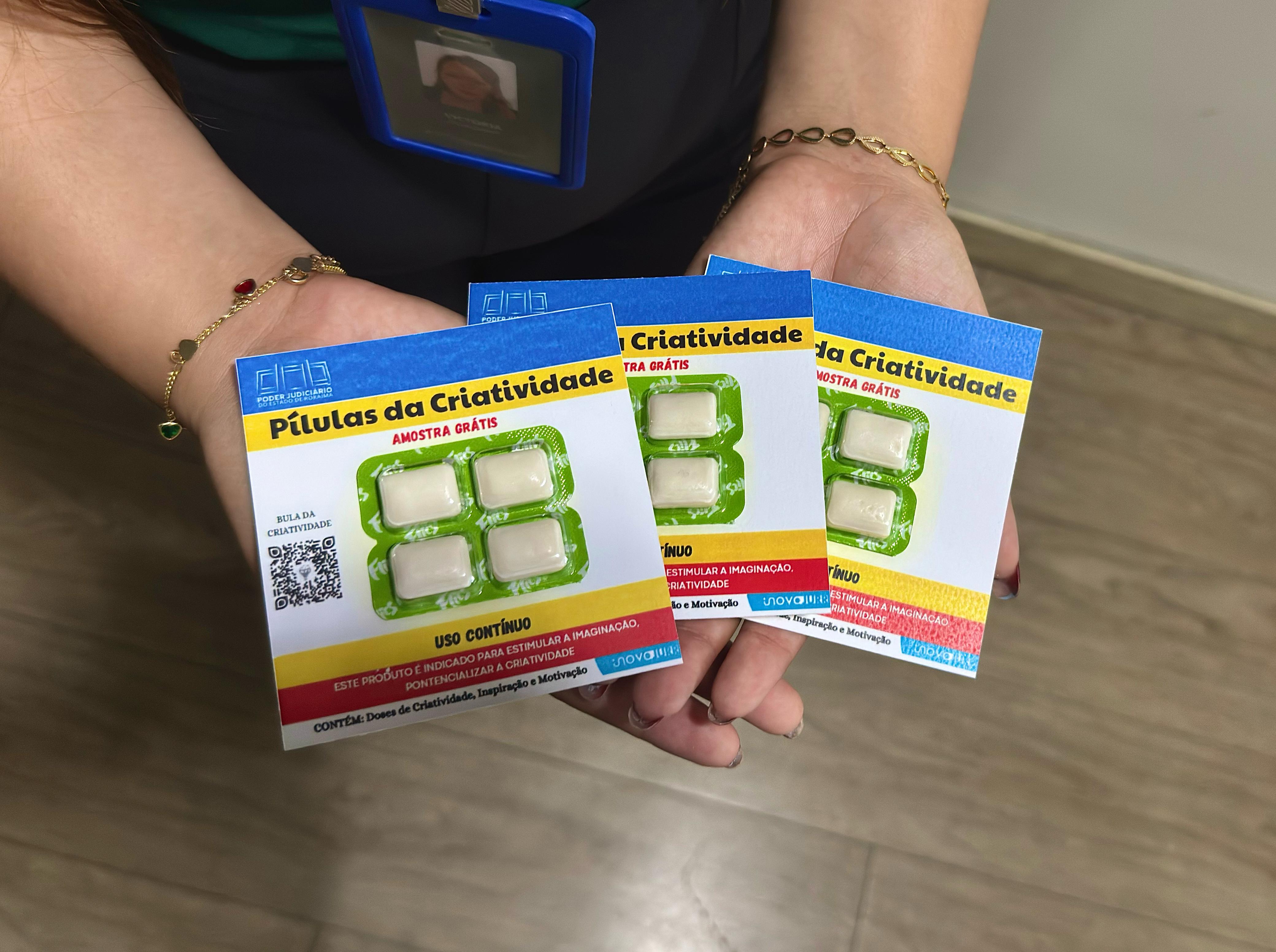 A imagem mostra as mãos de uma pessoa segurando três embalagens individuais de um produto chamado "Pílulas da Criatividade". Cada embalagem contém quatro pastilhas brancas, semelhantes a chicletes ou balas pequenas. A imagem mostra as mãos de uma pessoa segurando três embalagens individuais de um produto chamado "Pílulas da Criatividade". Cada embalagem contém quatro pastilhas brancas, semelhantes a chicletes ou balas pequenas.