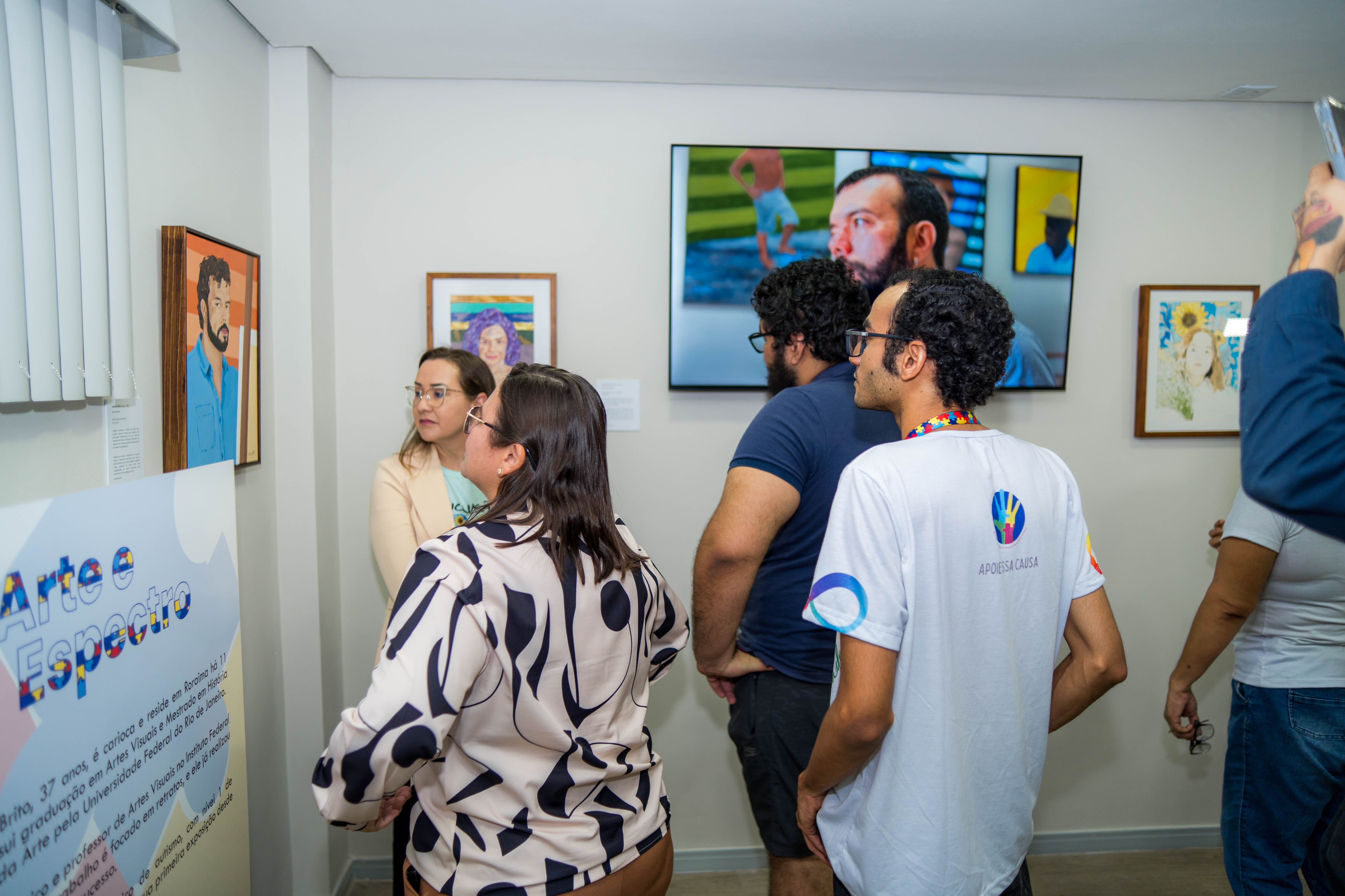 A imagem mostra um grupo de pessoas visitando uma exposição de arte. Eles estão em uma sala com paredes brancas e iluminação artificial. Há diversas obras de arte expostas nas paredes. A imagem mostra um grupo de pessoas visitando uma exposição de arte. Eles estão em uma sala com paredes brancas e iluminação artificial. Há diversas obras de arte expostas nas paredes.