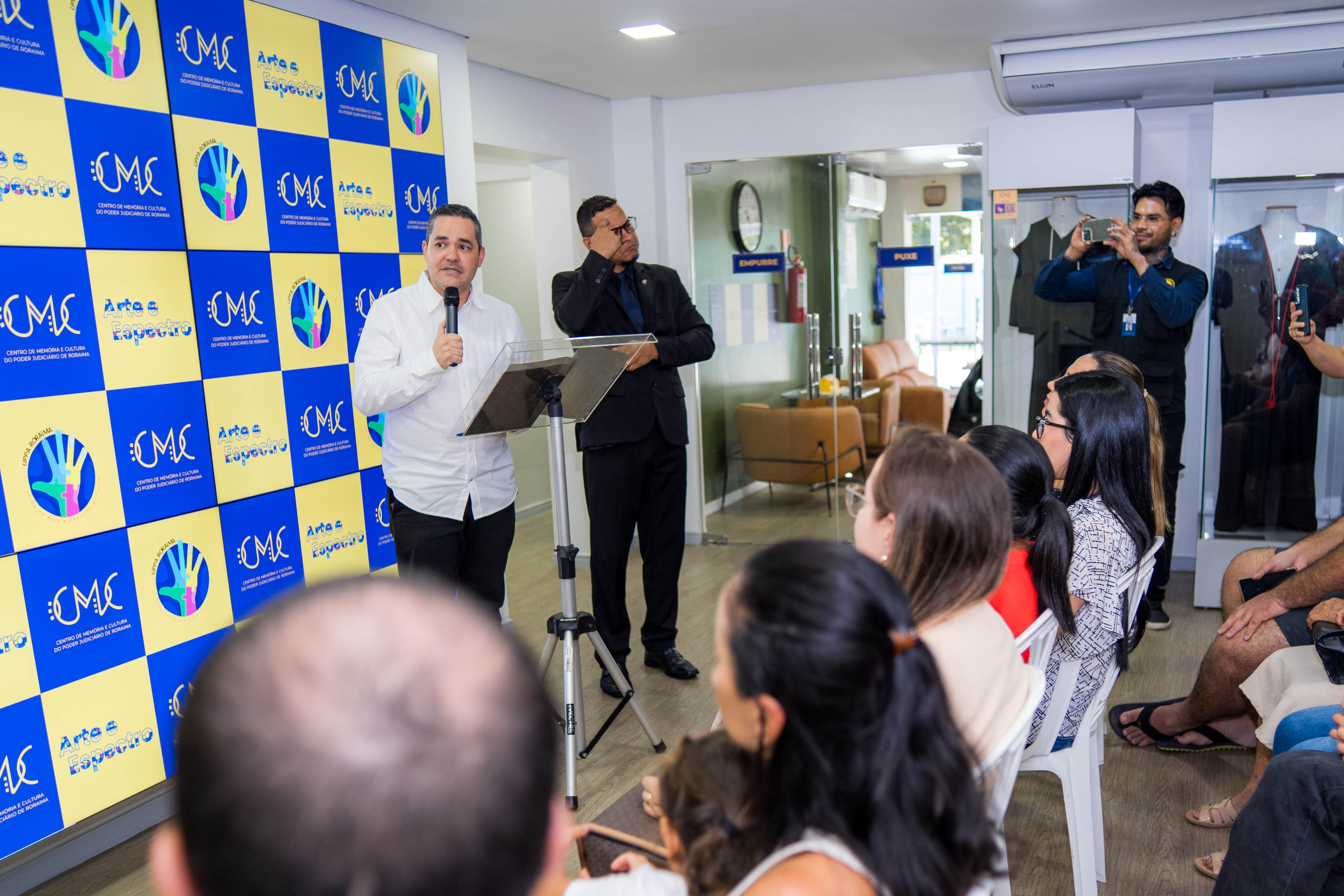A imagem captura o evento de inicio da exposição. No centro da imagem um homem de camisa branca e calças pretas está falando ao microfone em um púlpito com um tripé. A frente dele algumas pessoas sentadas assistem. Ao seu lado um interprete de libras. A imagem captura o evento de inicio da exposição. No centro da imagem um homem de camisa branca e calças pretas está falando ao microfone em um púlpito com um tripé. A frente dele algumas pessoas sentadas assistem. Ao seu lado um interprete de libras.