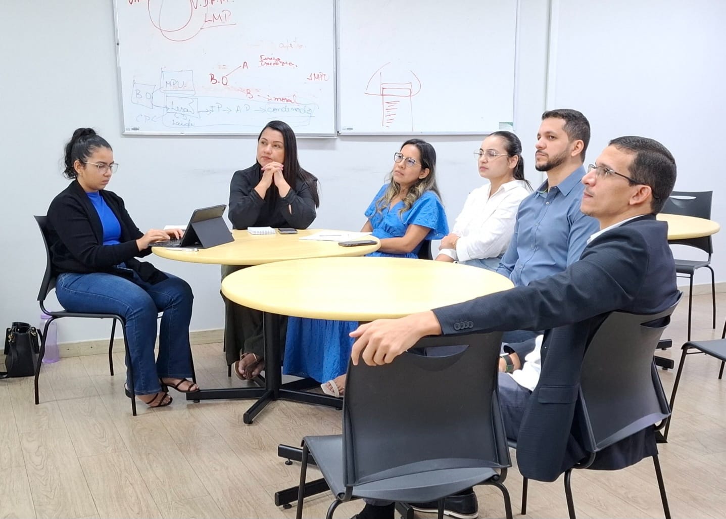 Imagem colorida contém seis pessoas, dois homens e quatro mulheres, ao redor de duas mesas circulares dentro de uma sala de aula. À esquerda, uma mulher manuseia um notebook. Ao centro, em primeiro plano, há uma cadeira vazia. Imagem colorida contém seis pessoas, dois homens e quatro mulheres, ao redor de duas mesas circulares dentro de uma sala de aula. À esquerda, uma mulher manuseia um notebook. Ao centro, em primeiro plano, há uma cadeira vazia.