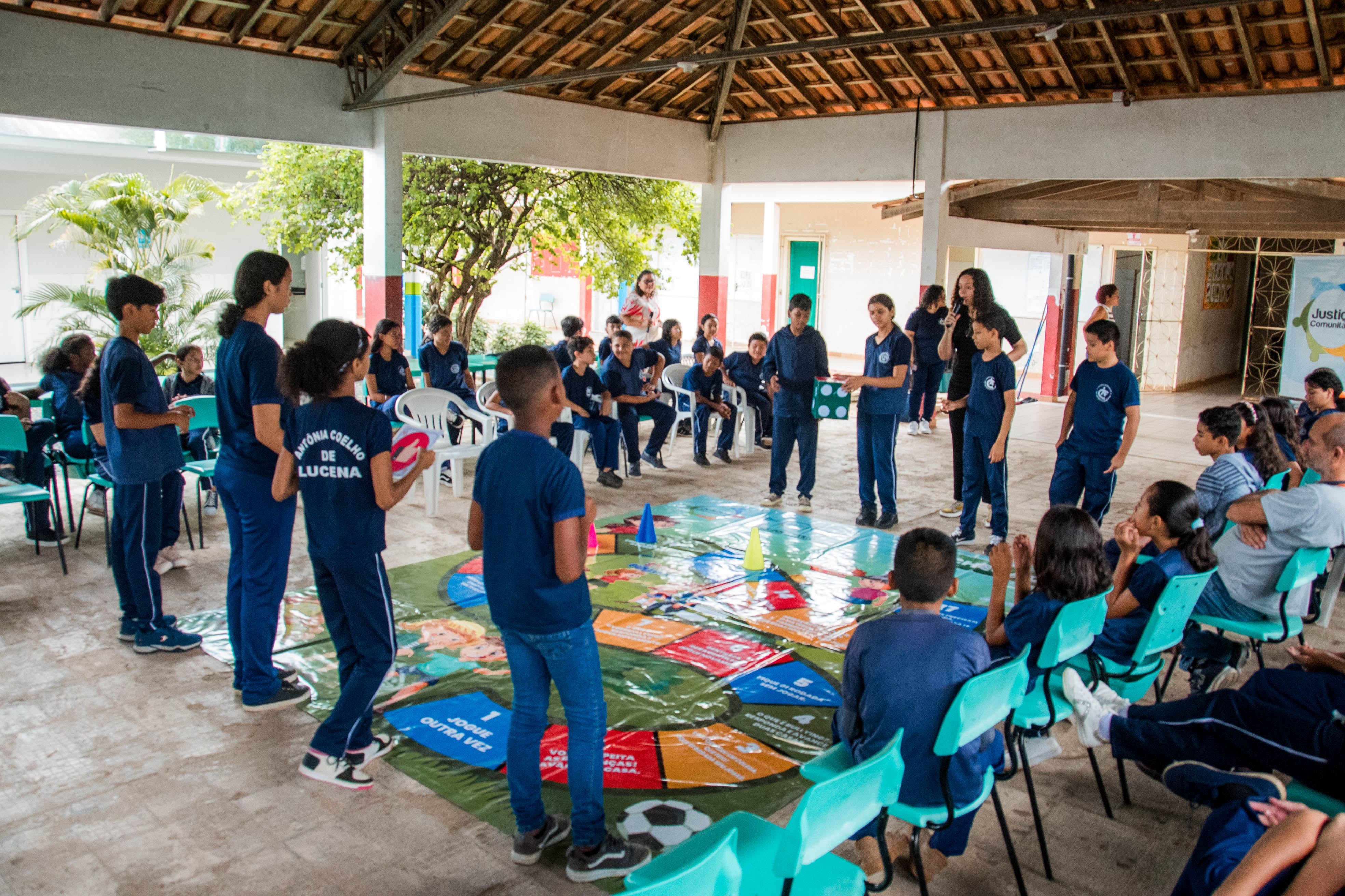 Alunos participam ativamente de uma dinâmica utilizando o tabuleiro gigante disposto no chão. Eles jogam um dado grande e interagem em grupo, enquanto outros assistem sentados ao redor.