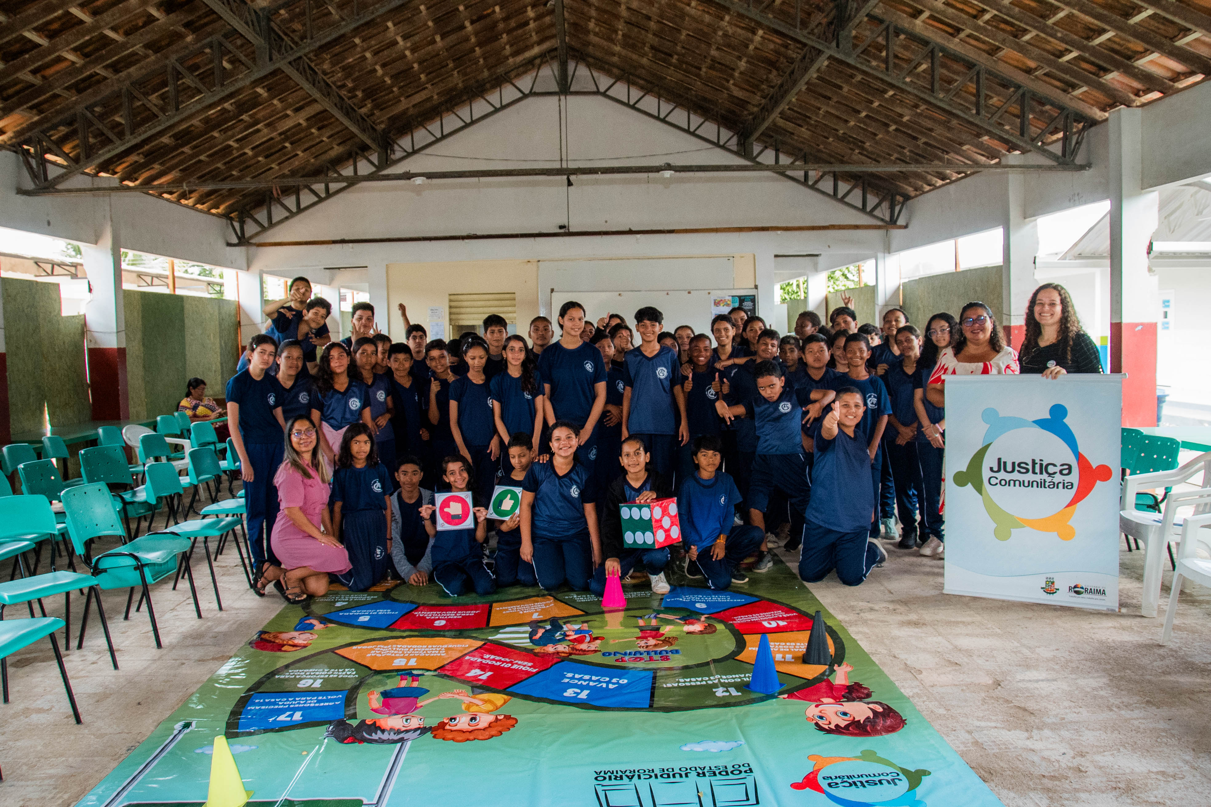 Um grupo de aproximadamente 49 alunos posa para uma foto em conjunto com as facilitadoras do projeto "Justiça Comunitária". Estão reunidos ao redor de um grande tabuleiro educativo no chão, que faz parte da atividade. Alguns estudantes seguram placas com símbolos de "curtir" e "não curtir", além de um dado gigante colorido. Um grupo de aproximadamente 49 alunos posa para uma foto em conjunto com as facilitadoras do projeto "Justiça Comunitária". Estão reunidos ao redor de um grande tabuleiro educativo no chão, que faz parte da atividade. Alguns estudantes seguram placas com símbolos de "curtir" e "não curtir", além de um dado gigante colorido.