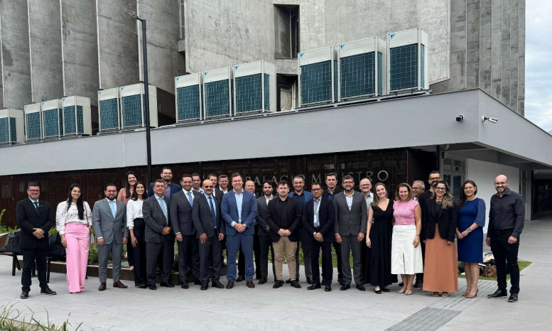 Foto colorida mostra um grupo de cerca de 25 pessoas vestidas formalmente posando em frente a um edifício moderno com fachada de concreto e unidades de ar-condicionado visíveis no topo. O letreiro parcial indica o nome do local. O grupo exibe expressões variadas, e o céu está nublado. Foto colorida mostra um grupo de cerca de 25 pessoas vestidas formalmente posando em frente a um edifício moderno com fachada de concreto e unidades de ar-condicionado visíveis no topo. O letreiro parcial indica o nome do local. O grupo exibe expressões variadas, e o céu está nublado.