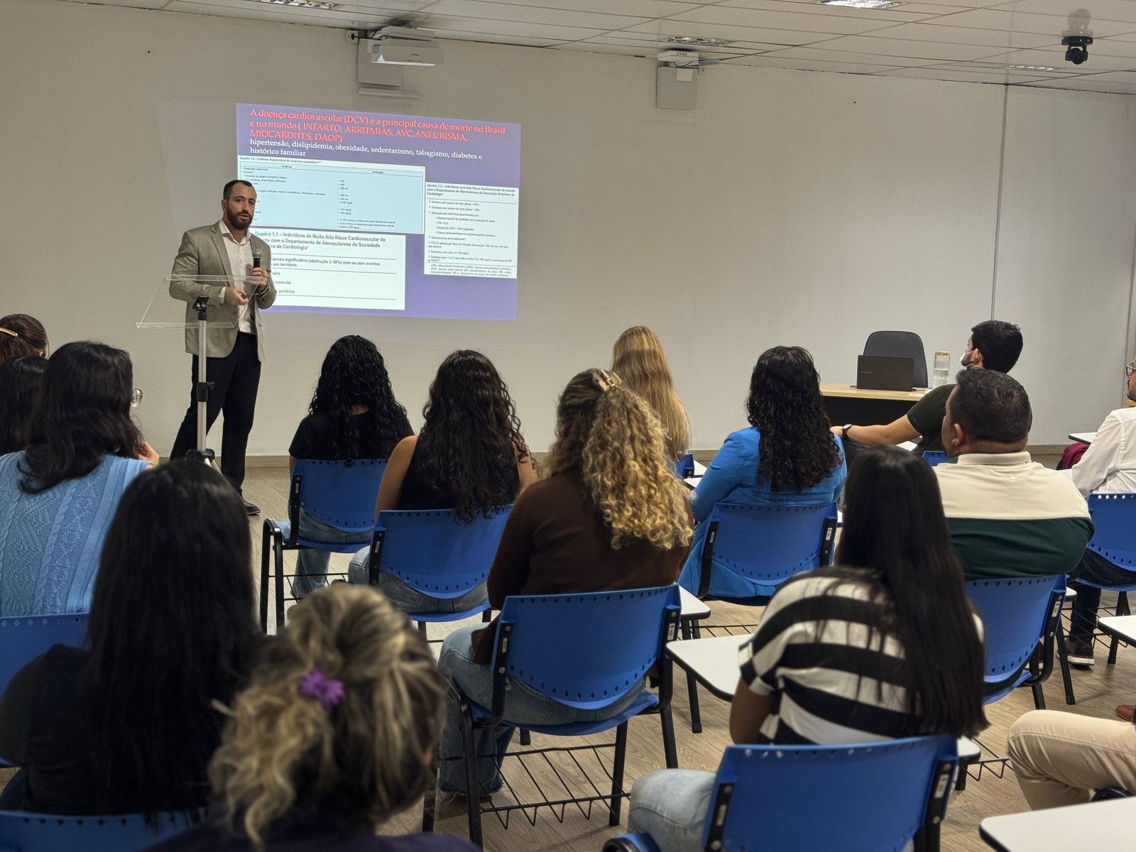 foto colorida de uma sala com pessoas sentadas em cadeira escolar enquanto o Secretário da Secretária de Qualidade de Vida, Hassan Syagha, está do lado do slide palestrando ao microfone. foto colorida de uma sala com pessoas sentadas em cadeira escolar enquanto o Secretário da Secretária de Qualidade de Vida, Hassan Syagha, está do lado do slide palestrando ao microfone.