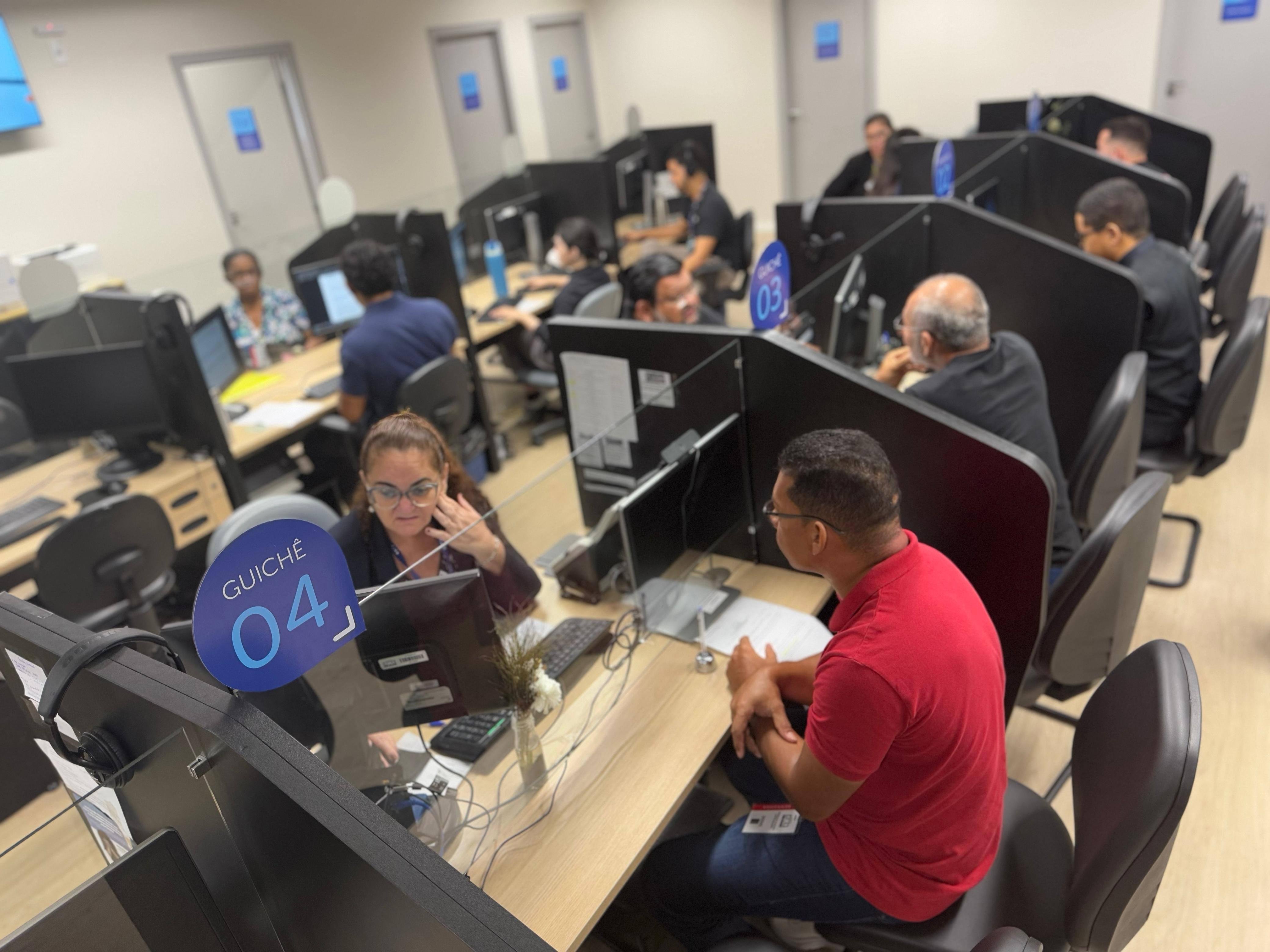A imagem retrata o interior de uma central de atendimento com estações de trabalho separadas por divisórias. Em destaque, uma mulher atende um homem no "Guichê 04", enquanto outras pessoas trabalham ao fundo. A imagem retrata o interior de uma central de atendimento com estações de trabalho separadas por divisórias. Em destaque, uma mulher atende um homem no "Guichê 04", enquanto outras pessoas trabalham ao fundo.