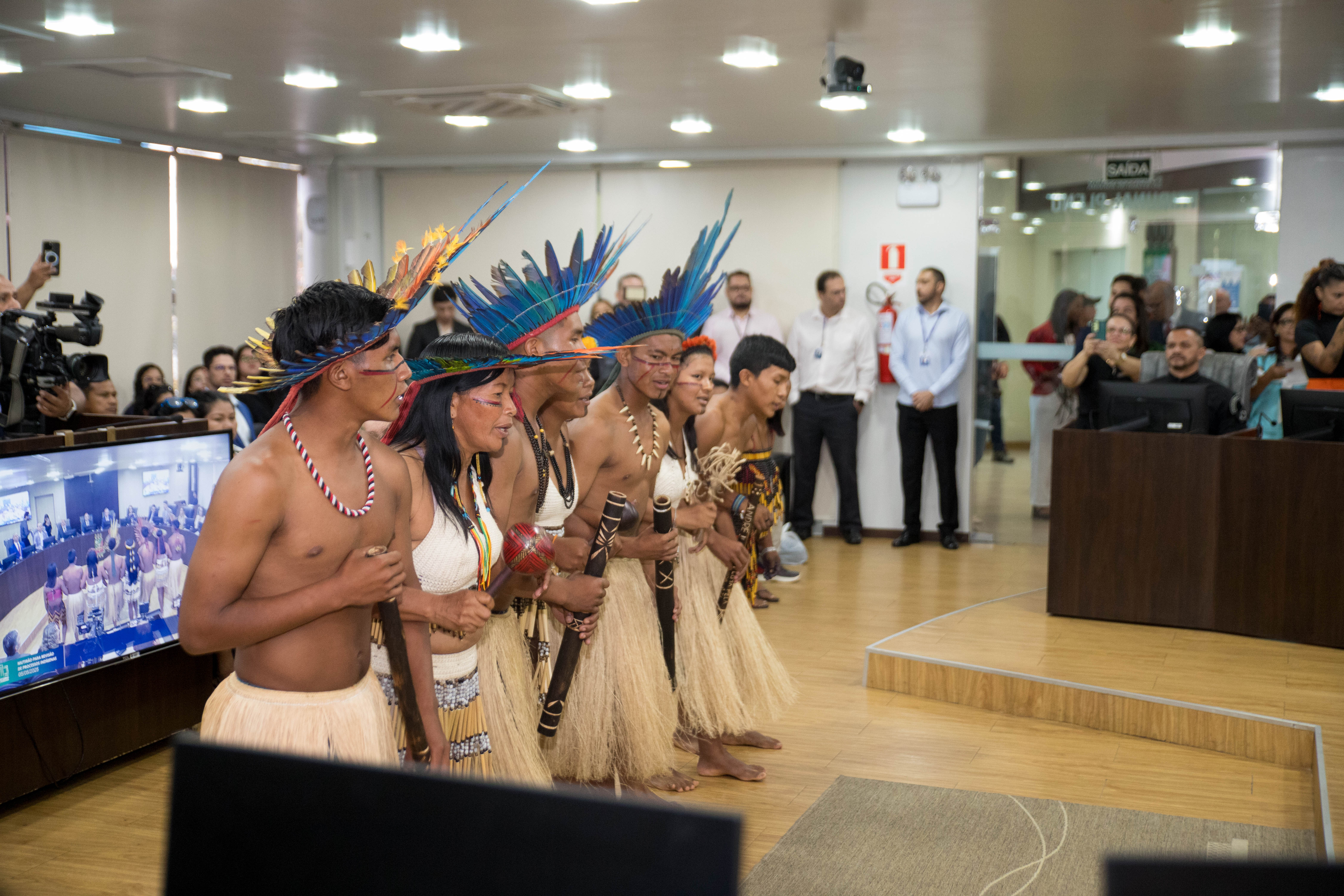 A imagem retrata um grupo de pessoas indígenas na Sala de Sessões do Tribunal Pleno realizando uma apresentação cultural. 
