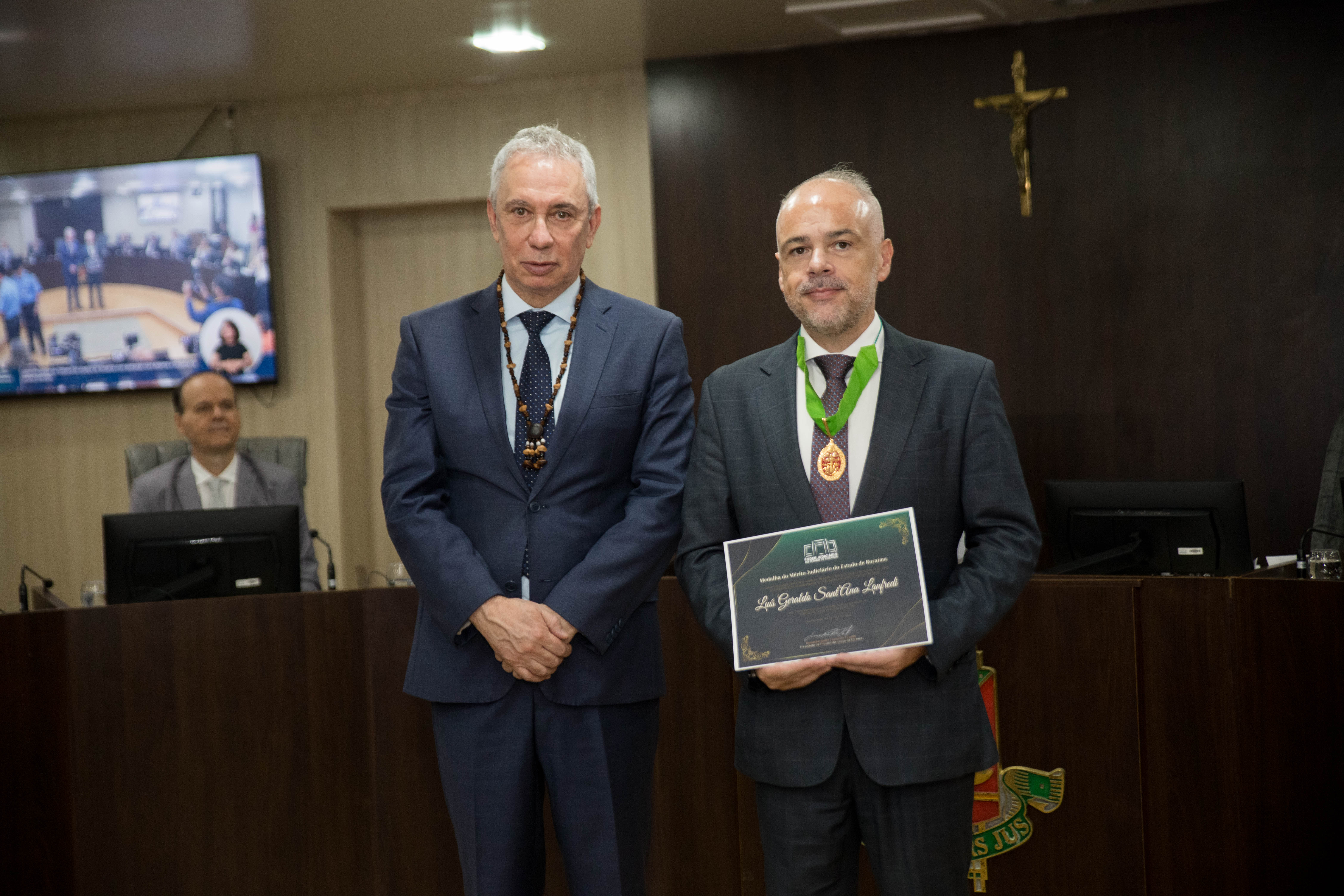 A imagem colorida mostra o Desembargador Luís Lanfredi segurando a Medalha do Mérito Judiciário do Estado de Roraima ao lado do vice-presidente do Tribunal de Justiça de Roraima e coordenador do GMF, Desembargador Almiro Padilha, 