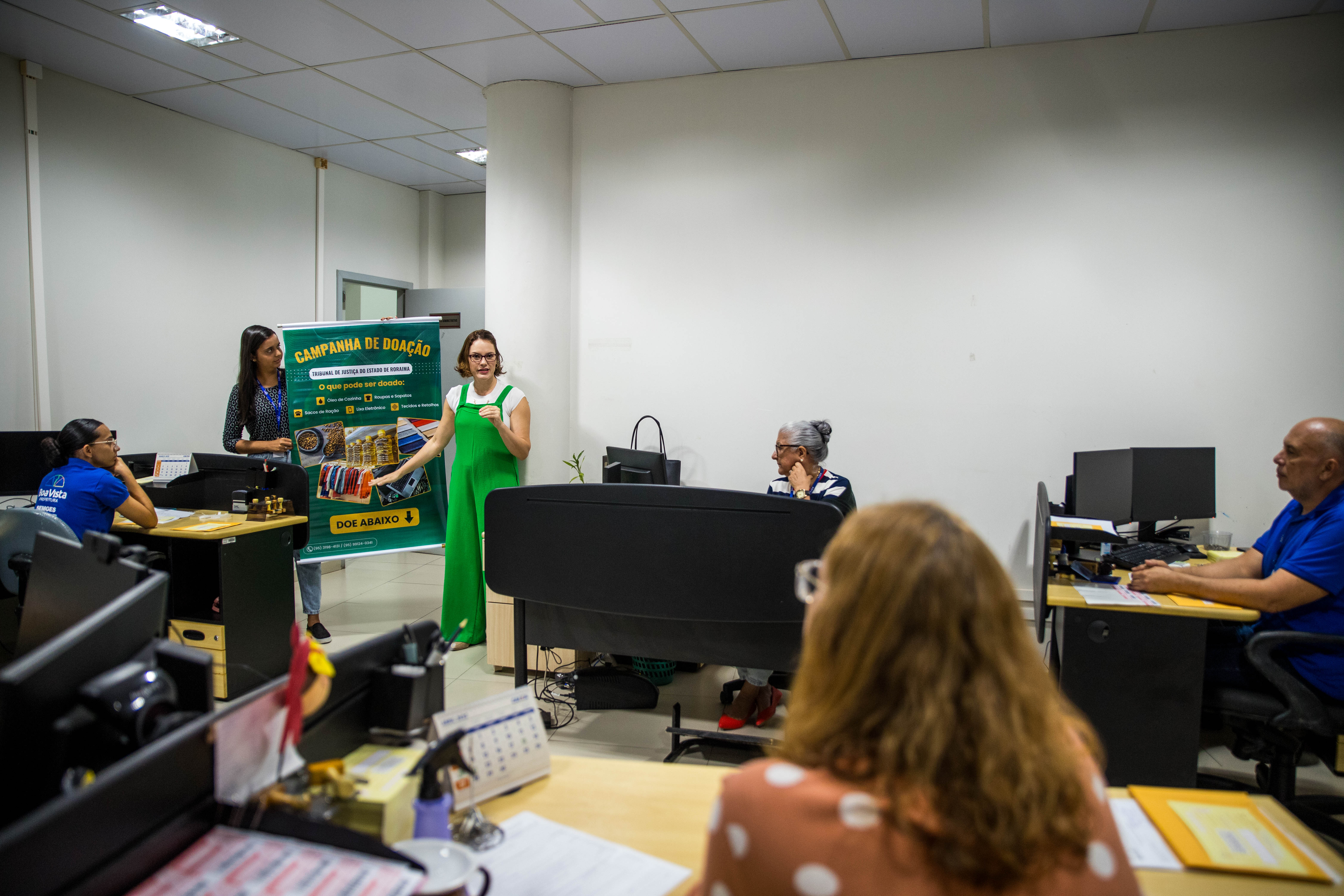 Foto colorida mostra uma mulher de macacão verde em um ambiente de escritório, no qual a mesma está apresentando uma campanha de doação para um grupo de funcionários. Ela está ao lado de um banner verde com informações sobre os itens que podem ser doados, como roupas, alimentos e livros. Algumas pessoas estão sentadas em suas mesas, ouvindo a apresentação, enquanto outra mulher segura o banner.