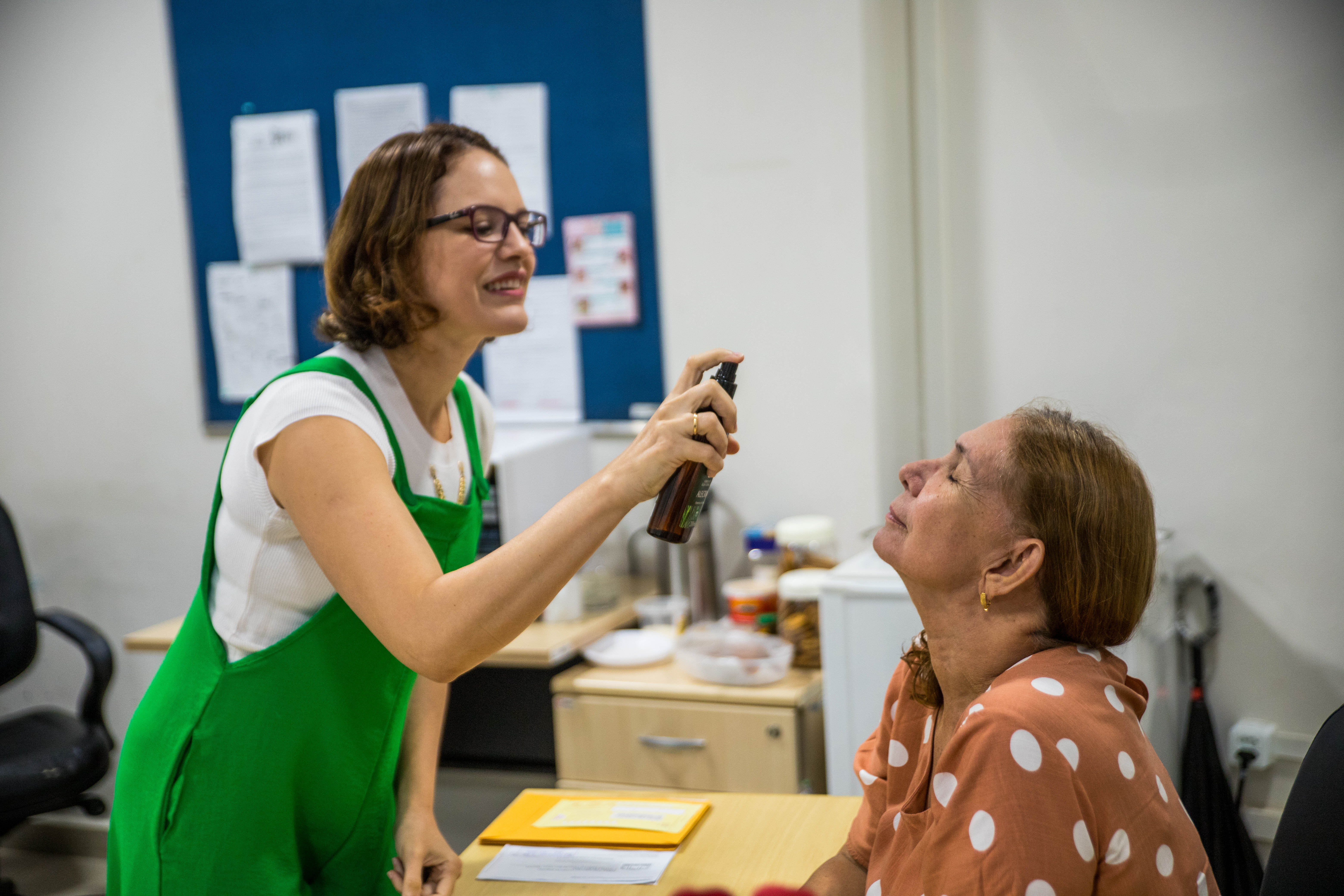 Foto colorida mostra uma mulher de macacão verde em um ambiente de escritório, no qual a mesma está aplicando um spray facial em outra mulher que está sentada à mesa, de olhos fechados e expressão relaxada. O clima da imagem transmite um momento de descontração no ambiente de trabalho.