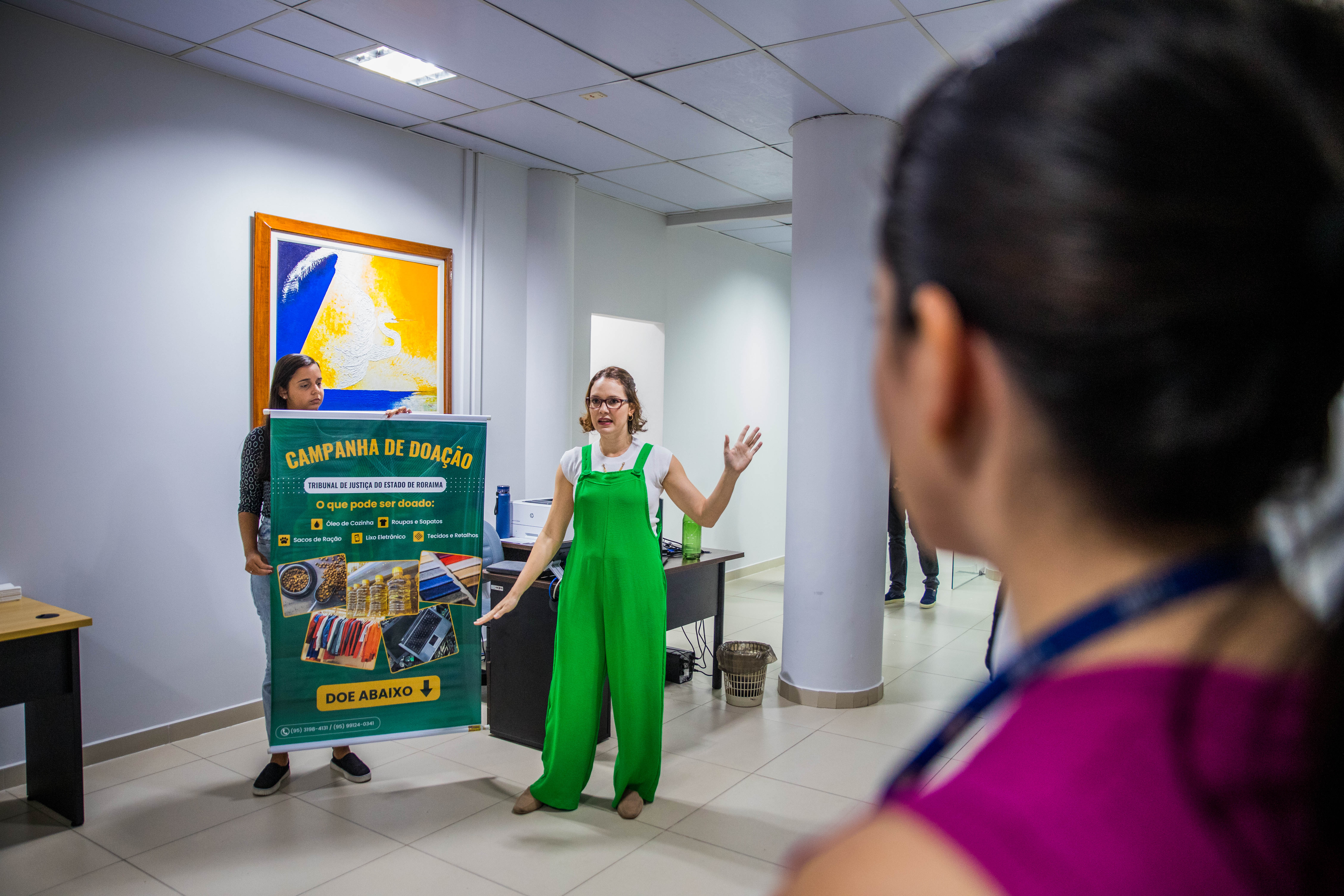 Foto colorida mostra a mesma mulher de macacão verde está explicando sobre a campanha de doação para um grupo de pessoas em um corredor do escritório. O banner da campanha está sendo segurado por outra mulher ao lado dela. Em primeiro plano, há uma pessoa de costas, ouvindo atentamente a explicação.