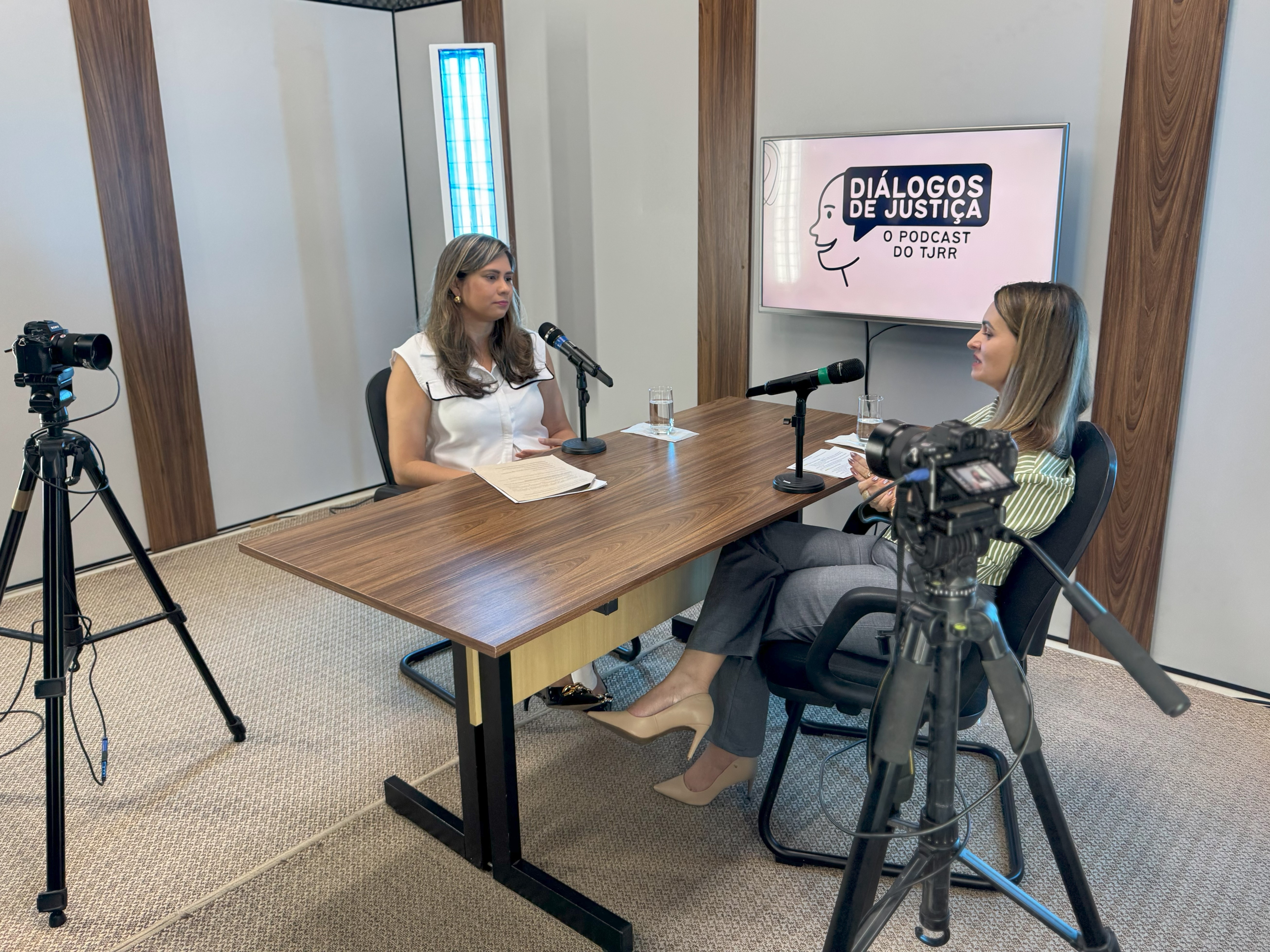 A imagem mostra um estúdio de gravação de podcast com duas mulheres sentadas frente a frente em uma mesa de madeira, cada uma com microfone e câmera profissional direcionados para si. A mulher à esquerda tem cabelos castanhos claros e veste blusa branca com detalhes pretos, enquanto a da direita tem cabelos loiros e usa blusa listrada em verde e branco, gesticulando durante a fala. Ao fundo, uma TV exibe o logotipo do podcast “Diálogos de Justiça – O podcast do TJRR”, indicando que o conteúdo é produzido pelo Tribunal de Justiça de Roraima. O estúdio possui iluminação suave, carpete em tons de bege e marrom e painéis acústicos para aprimorar a qualidade sonora. A imagem mostra um estúdio de gravação de podcast com duas mulheres sentadas frente a frente em uma mesa de madeira, cada uma com microfone e câmera profissional direcionados para si. A mulher à esquerda tem cabelos castanhos claros e veste blusa branca com detalhes pretos, enquanto a da direita tem cabelos loiros e usa blusa listrada em verde e branco, gesticulando durante a fala. Ao fundo, uma TV exibe o logotipo do podcast “Diálogos de Justiça – O podcast do TJRR”, indicando que o conteúdo é produzido pelo Tribunal de Justiça de Roraima. O estúdio possui iluminação suave, carpete em tons de bege e marrom e painéis acústicos para aprimorar a qualidade sonora.