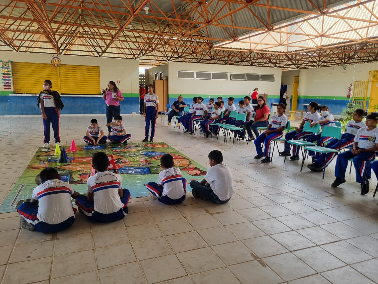 Imagem colorida mostra um pátio escolar coberto, com um telhado de treliça, onde um grupo de estudantes e alguns adultos estão reunidos. No centro, há um grande tapete de chão que se assemelha a um jogo de tabuleiro com ilustrações coloridas, e um cone amarelo e outro rosa estão sobre ele. Vários estudantes, vestindo uniformes azuis e brancos, estão sentados no chão ao redor do tapete, alguns de frente para a câmera e outros para o jogo. Outros estudantes e adultos estão sentados em cadeiras de plástico verdes e brancas, formando um semicírculo ao redor da área principal do jogo. Uma mulher, provavelmente uma professora, está em pé perto do tapete, segurando algo em suas mãos, e parece estar conduzindo a atividade. O ambiente é bem iluminado, e há paredes brancas e uma porta amarela fechada ao fundo. Imagem colorida mostra um pátio escolar coberto, com um telhado de treliça, onde um grupo de estudantes e alguns adultos estão reunidos. No centro, há um grande tapete de chão que se assemelha a um jogo de tabuleiro com ilustrações coloridas, e um cone amarelo e outro rosa estão sobre ele. Vários estudantes, vestindo uniformes azuis e brancos, estão sentados no chão ao redor do tapete, alguns de frente para a câmera e outros para o jogo. Outros estudantes e adultos estão sentados em cadeiras de plástico verdes e brancas, formando um semicírculo ao redor da área principal do jogo. Uma mulher, provavelmente uma professora, está em pé perto do tapete, segurando algo em suas mãos, e parece estar conduzindo a atividade. O ambiente é bem iluminado, e há paredes brancas e uma porta amarela fechada ao fundo.