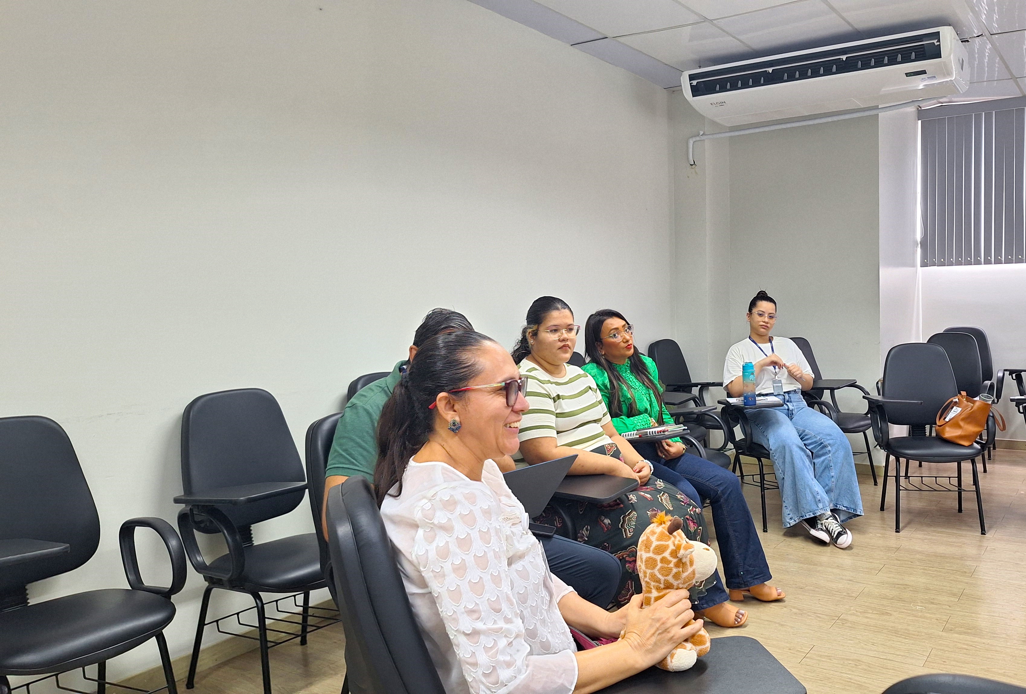  foto colorida dando enfoque no meio circulo de participantes. Em primeiro plano está uma participante sorrindo enquanto segura uma girafa de pelúcia.