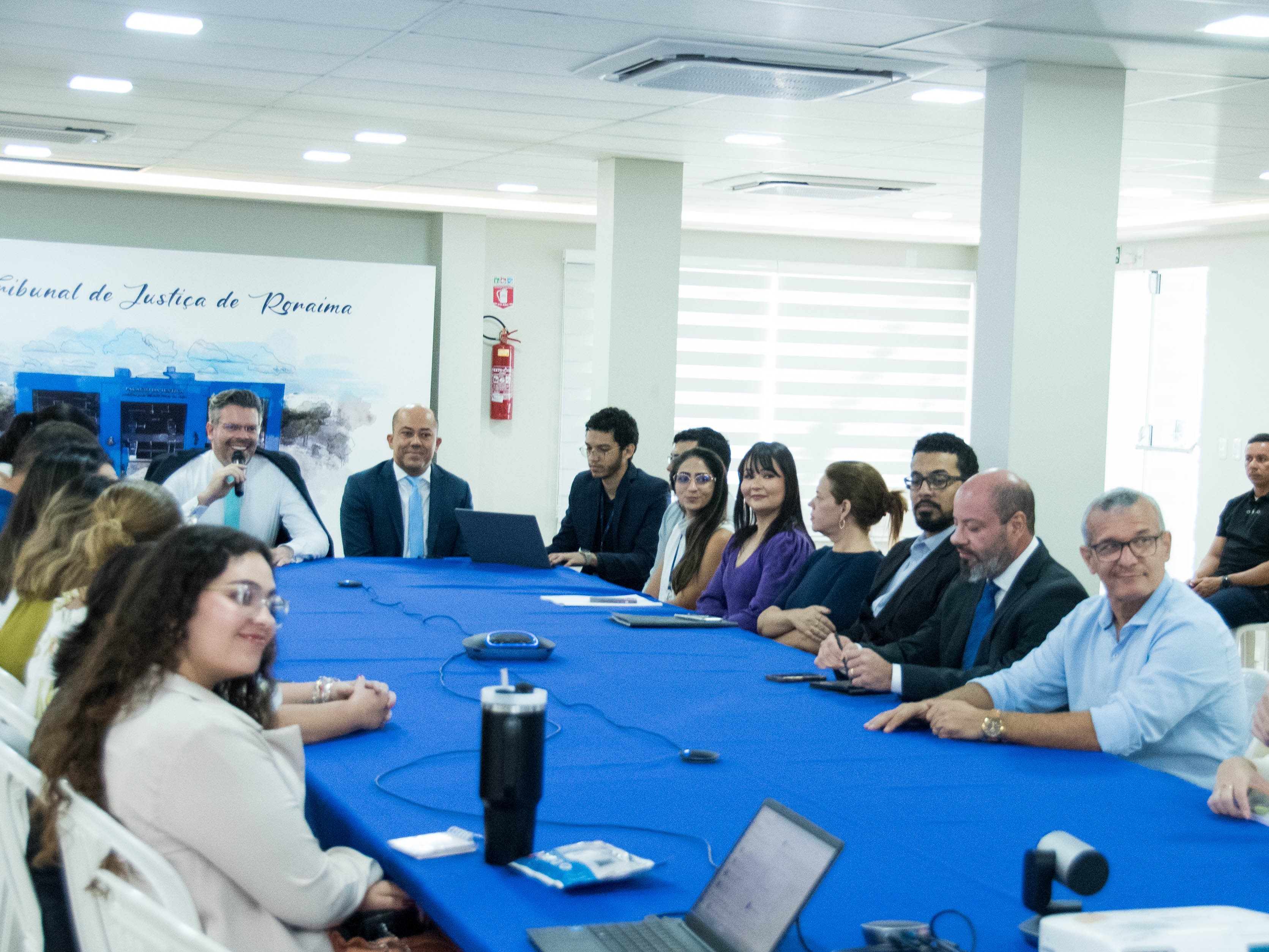 Foto colorida mostra um grupo de 15 pessoas sentadas em uma mesa de conferência azul, participando de uma reunião. Foto colorida mostra um grupo de 15 pessoas sentadas em uma mesa de conferência azul, participando de uma reunião.