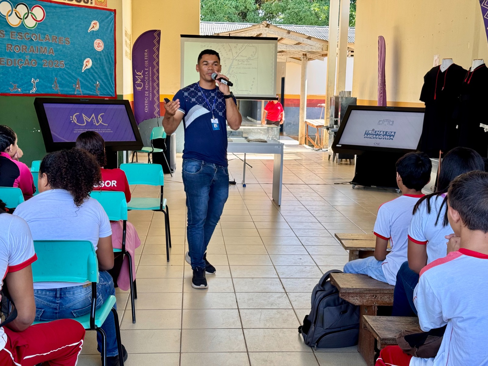 A imagem colorida mostra um homem em pé, no centro, vestindo uma camiseta azul e calça jeans, segurando um microfone e gesticulando enquanto fala para um grupo de jovens sentados em cadeiras e bancos à sua frente. Ao fundo, há um telão com projeção, e à direita e à esquerda, painéis digitais. O ambiente é um espaço aberto, parcialmente coberto, que parece ser uma área de eventos ou pátio escolar, com árvores e construções ao fundo. No lado esquerdo, um banner azul com o logotipo olímpico e o texto "Jogos Escolares de Roraima Edição 2025" é visível.