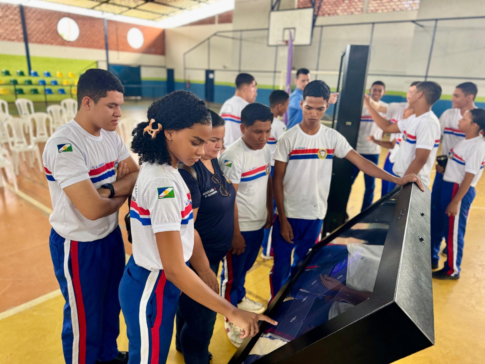 A imagem colorida mostra um grupo de jovens, a maioria vestindo uniformes de camiseta branca e calça azul com listras laterais vermelhas e brancas, reunidos ao redor de uma tela interativa inclinada, de cor escura. Uma jovem no centro da imagem está apontando para a tela, enquanto os outros a observam. O ambiente é interno, possivelmente um ginásio ou salão escolar, com o chão em tons de laranja e algumas cadeiras brancas ao fundo.
