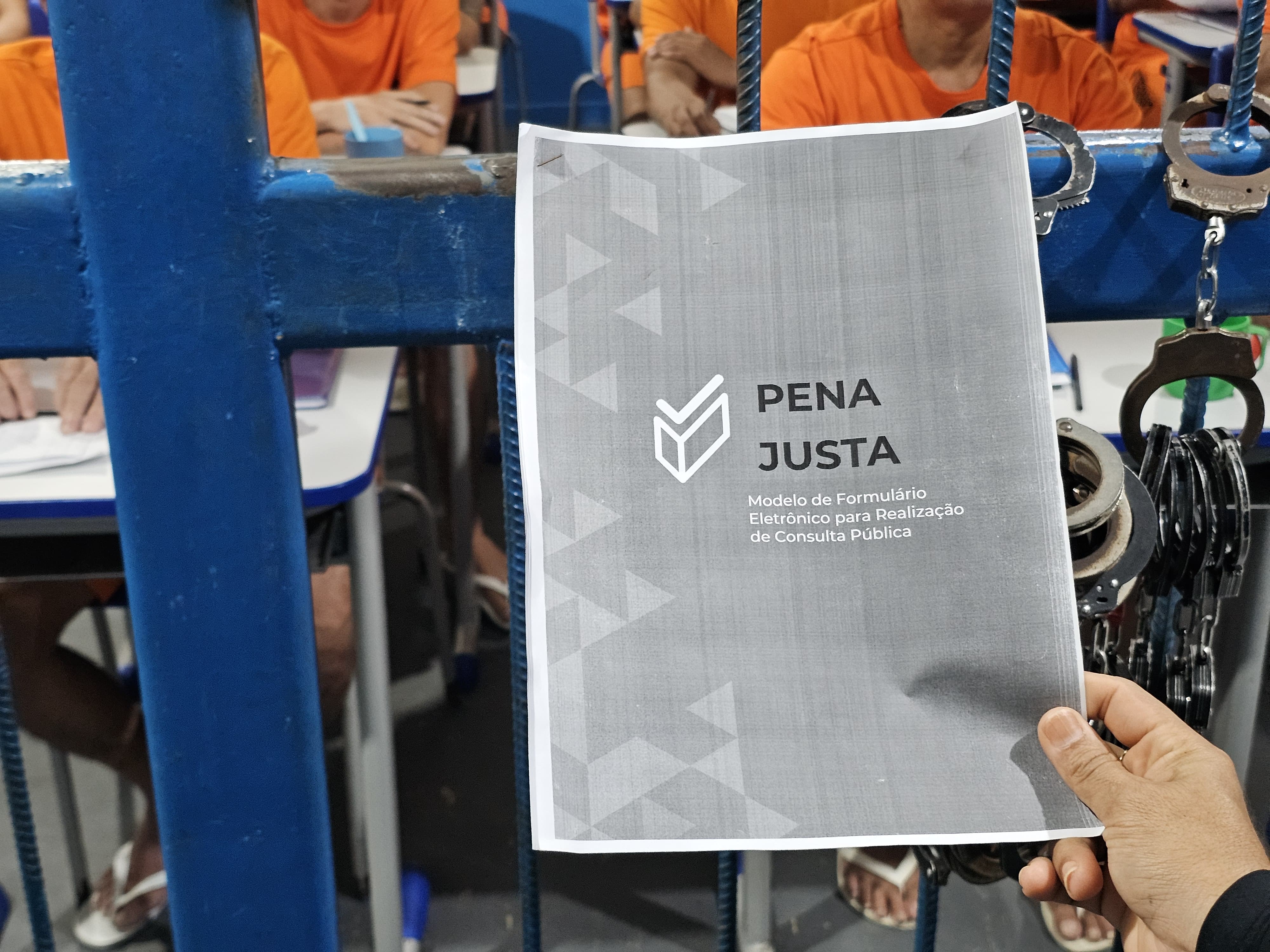Foto colorida mostra a capa do formulário “PENA JUSTA – Modelo de Formulário Eletrônico para Realização de Consulta Pública”, segurado por uma pessoa. Ao fundo, reeducandos com camisetas laranja estão sentados Foto colorida mostra a capa do formulário “PENA JUSTA – Modelo de Formulário Eletrônico para Realização de Consulta Pública”, segurado por uma pessoa. Ao fundo, reeducandos com camisetas laranja estão sentados